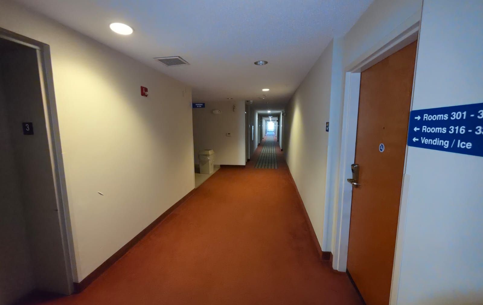 Un pasillo de hotel con piso alfombrado, puertas a la izquierda y un cartel que indica las habitaciones 301-303 y máquinas expendedoras/hielo a la derecha.