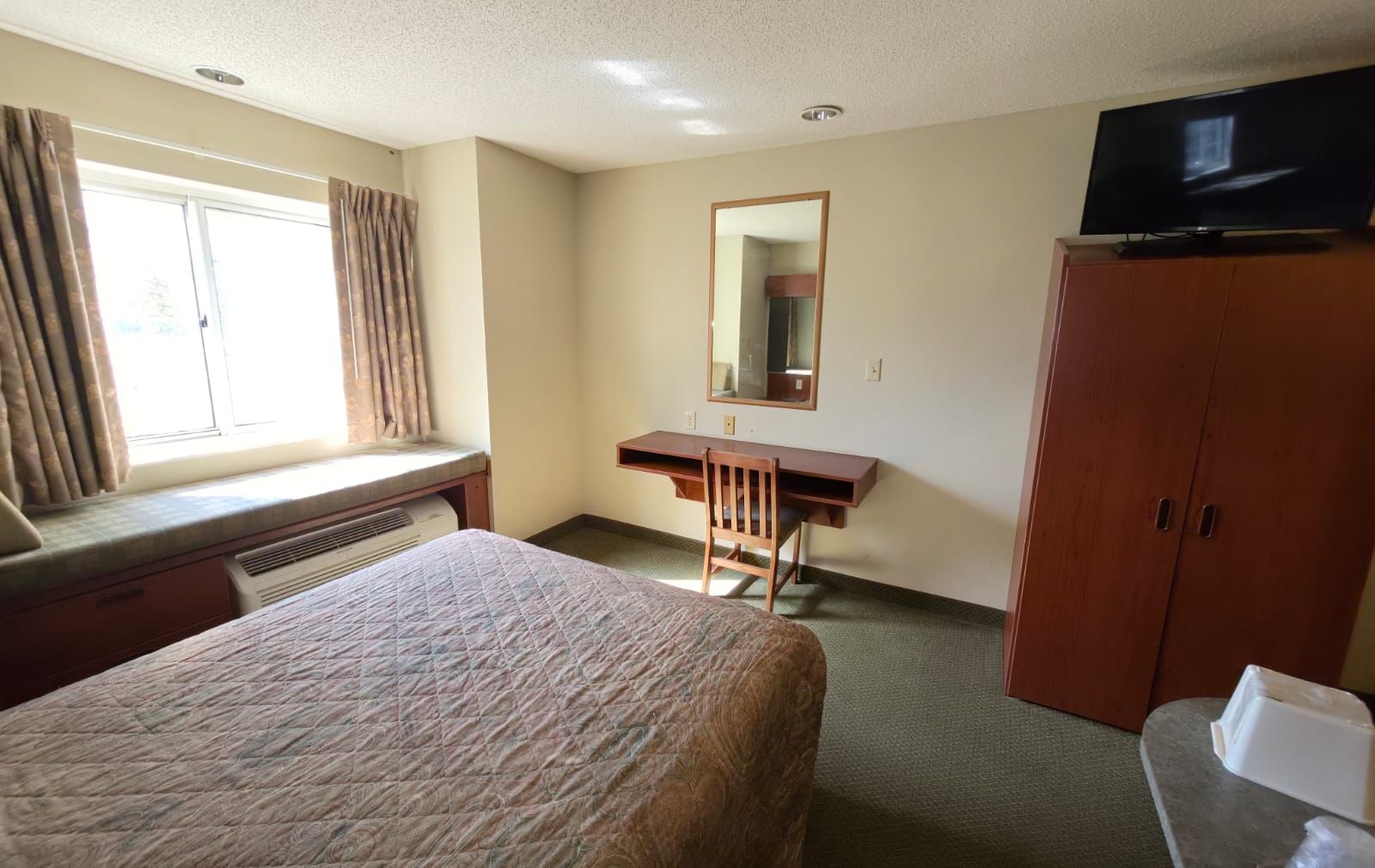 Habitación de hotel con cama, escritorio, silla, televisor, armario y ventana con cortinas.
