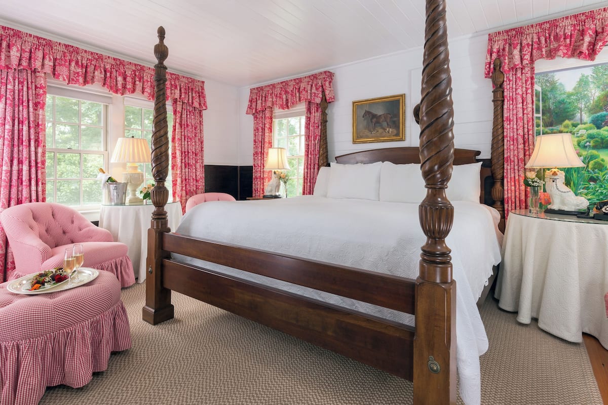 A four-poster bed with a white bedspread, two bedside tables with lamps, a chair with a tray of food, and a window with a view of greenery.