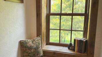 A wooden bench with a cushion, a window with a view of trees, and a small shelf with books and a figurine.