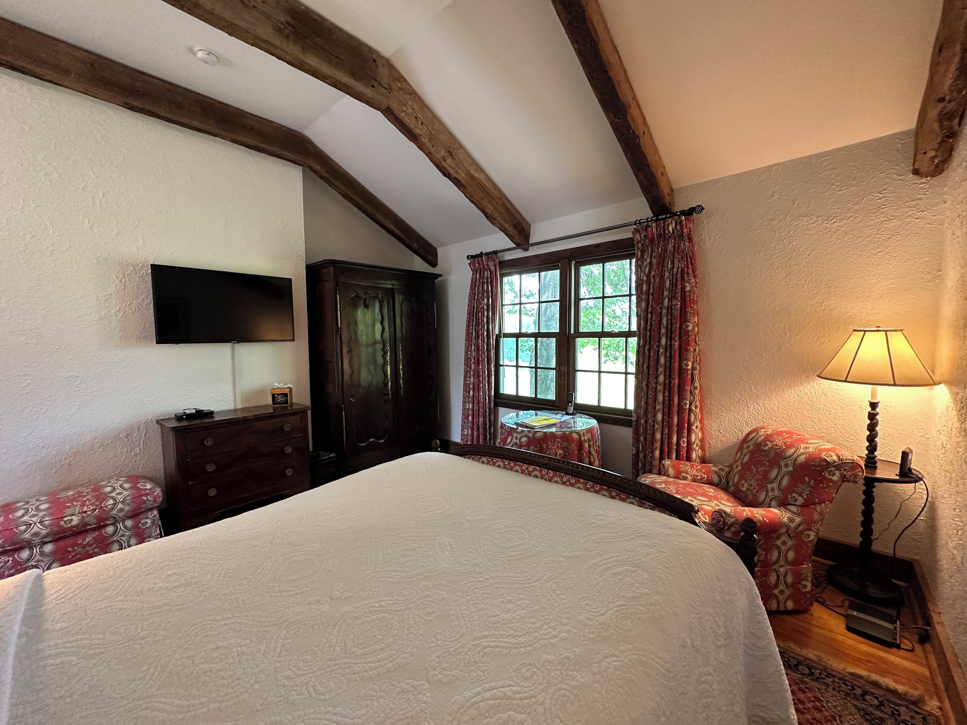 A bedroom with a large bed, wooden beams, a wardrobe, a desk, a chair, a lamp, and a window with curtains.