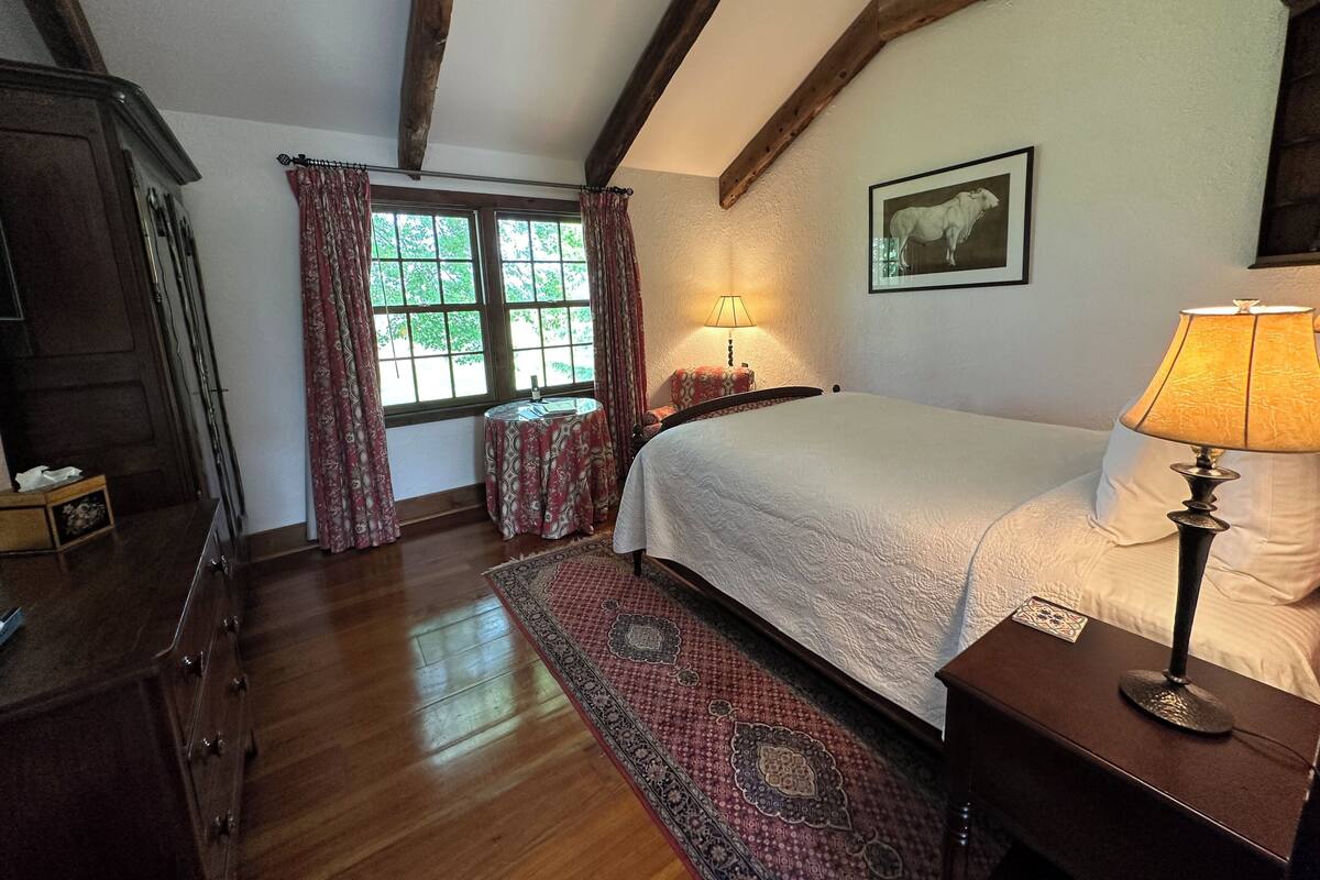 A bedroom with a wooden floor, a bed with a white duvet, a nightstand with a lamp, a window with curtains, and a framed picture on the wall.