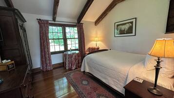 A bedroom with a wooden floor, a bed with a white duvet, a nightstand with a lamp, a window with curtains, and a framed picture on the wall.