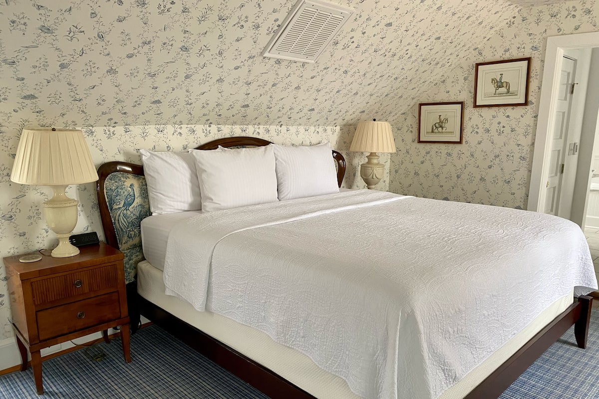 A neatly made bed with a white bedspread, two bedside lamps, a wooden nightstand with a drawer, and floral wallpaper.