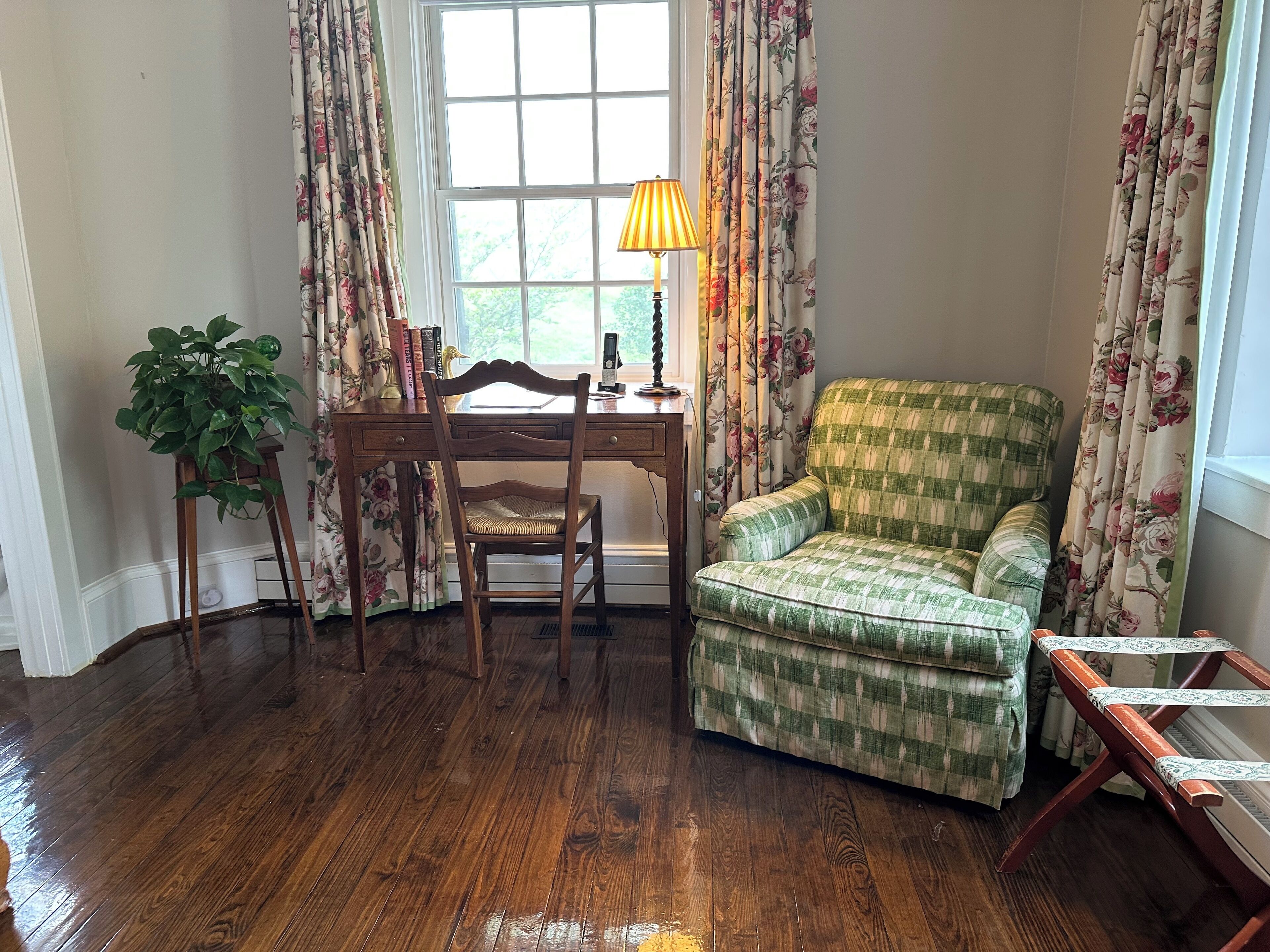 Una habitación con un escritorio de madera, un sillón verde a cuadros, una planta en maceta y una ventana con cortinas estampadas de flores.