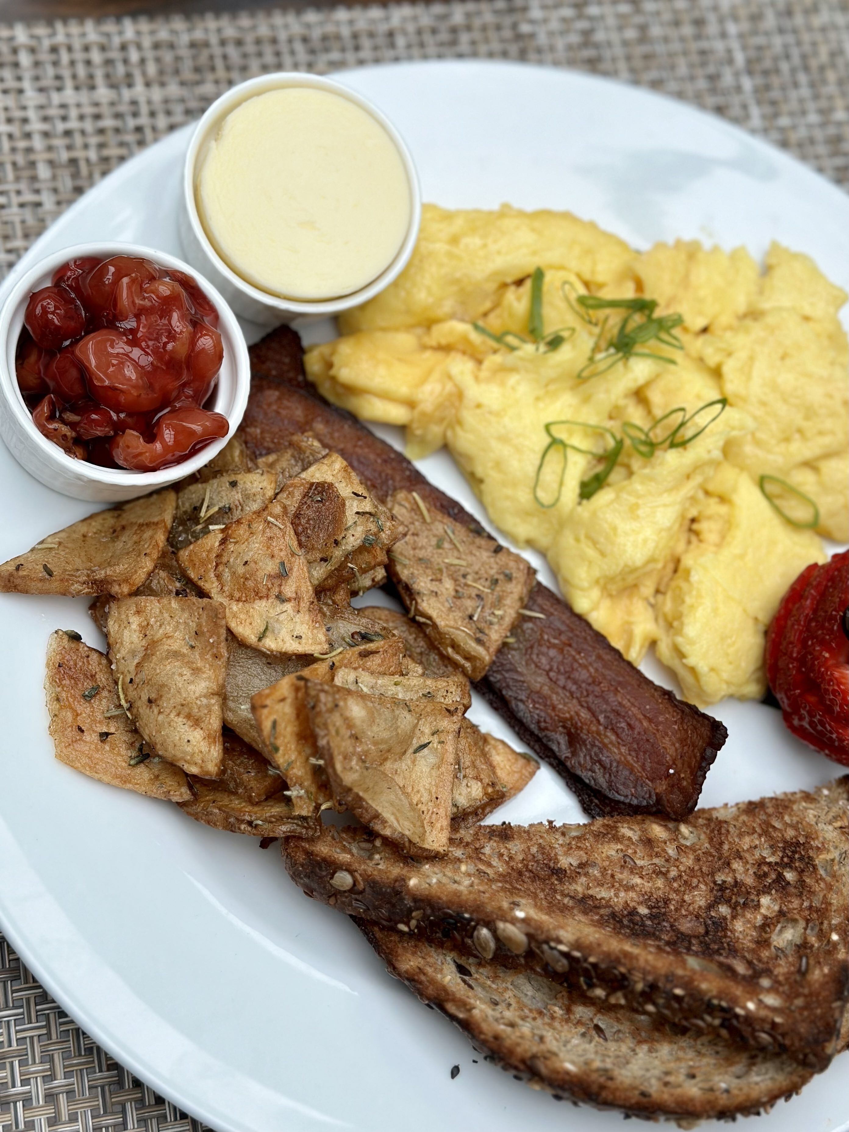 Un plato con huevos revueltos, tocino, papas asadas y un tazón pequeño de kétchup.