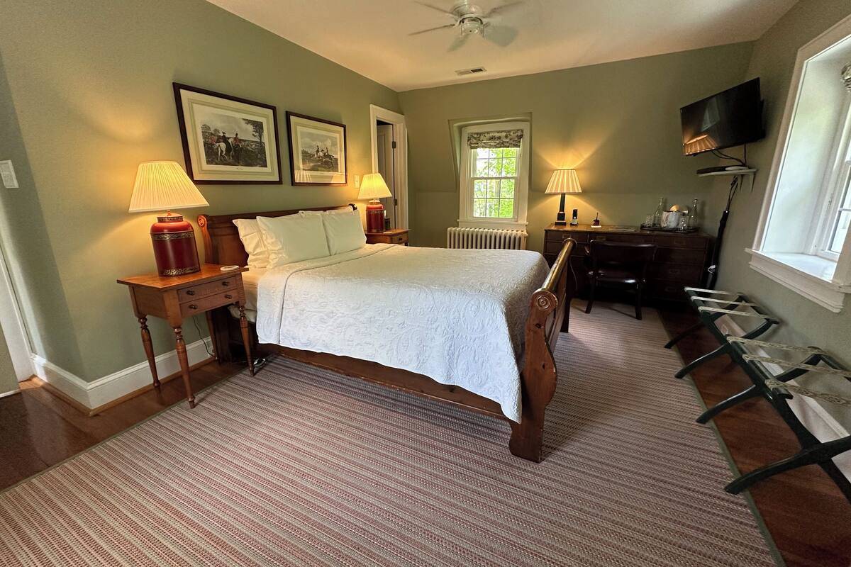 A bedroom with a bed, two bedside tables with lamps, a dresser, a chair, a TV, and a window with a view of greenery.
