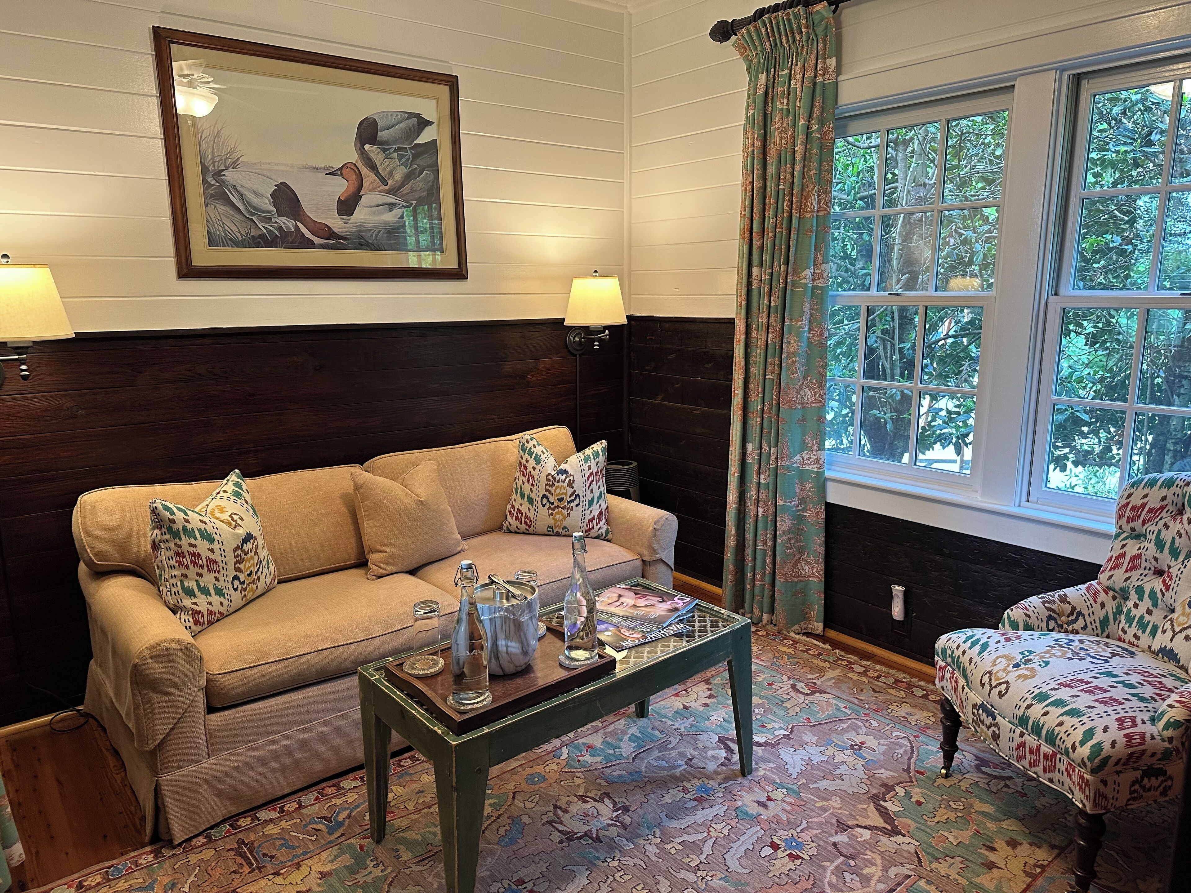 A cozy room with a beige sofa, a patterned armchair, a wooden coffee table, and a large window with curtains.