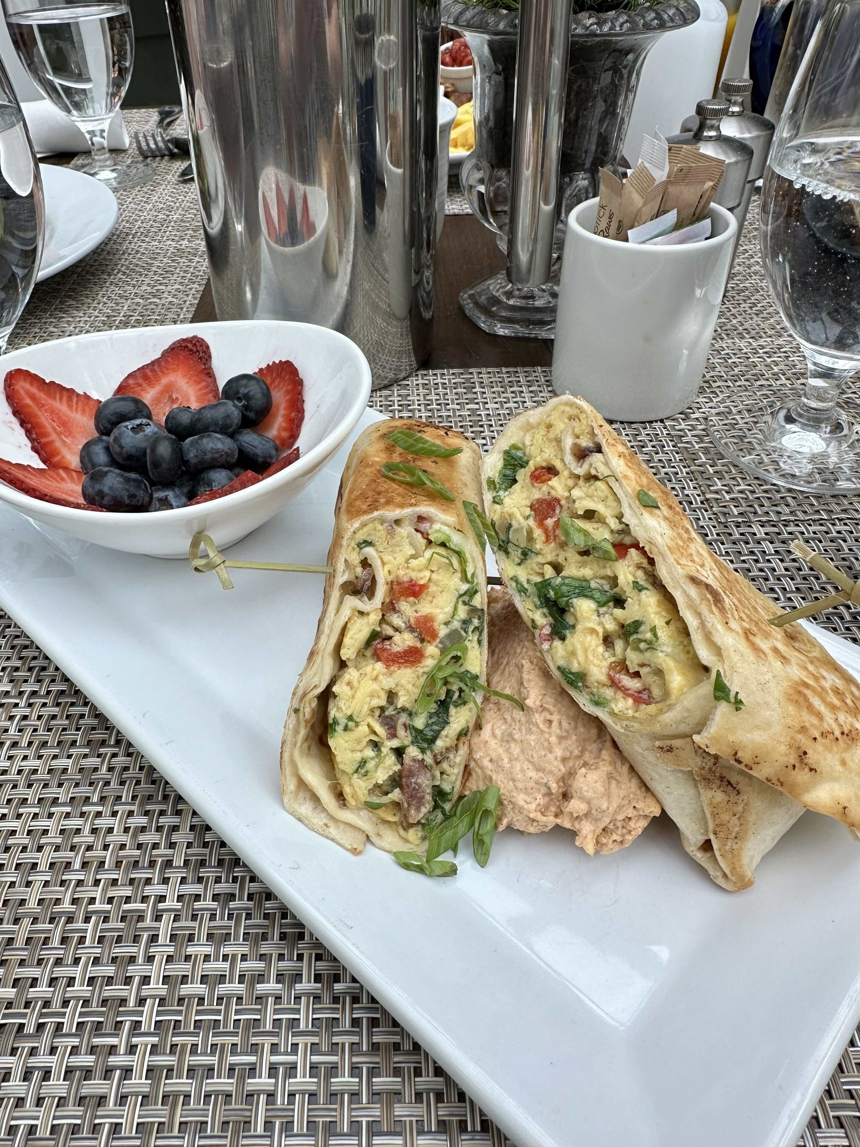 Une assiette de petit-déjeuner avec un sandwich enroulé, un bol de fraises et de myrtilles, et un verre d’eau sur une table en métal.