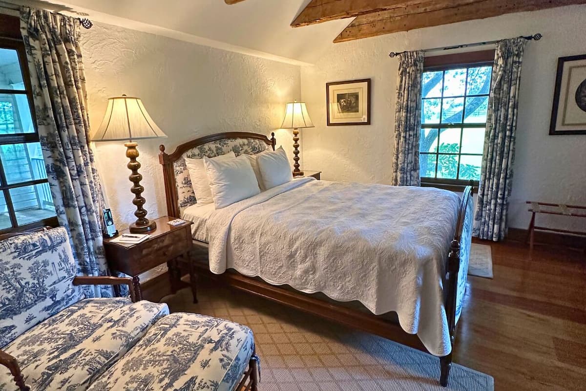 A bedroom with a large bed, a wooden nightstand, a chair, and two windows with curtains.