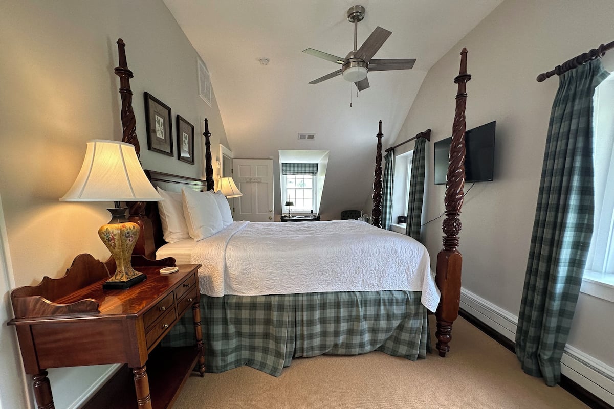 A bedroom with a four-poster bed, a ceiling fan, a television mounted on the wall, and a window with curtains.