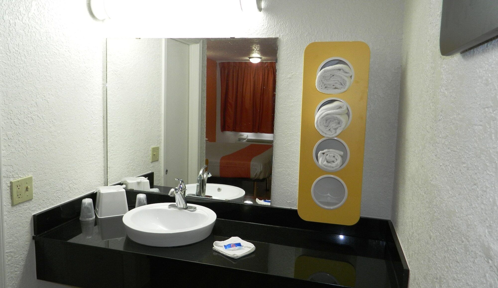 Un baño de hotel con un lavamanos blanco, encimera negra y un espejo con un sujetapapeles amarillo.