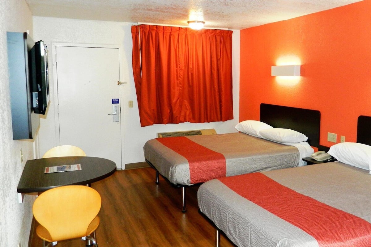 Habitación de hotel con dos camas, un escritorio, una silla, un televisor y una ventana con cortinas rojas.