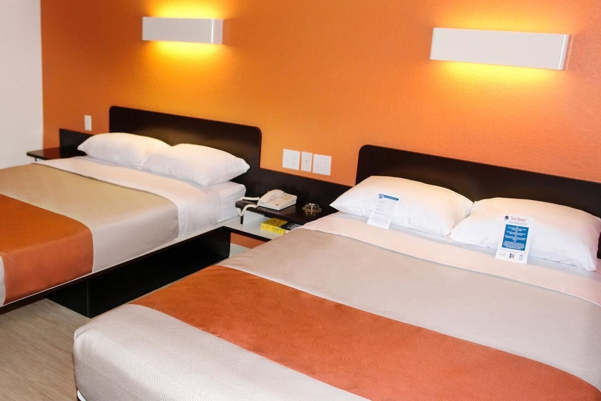 Habitación de hotel con dos camas, cada una con un almohadón blanco y un detalle marrón. Hay lámparas de pared sobre las camas y una mesita de noche con un teléfono y un objeto pequeño.