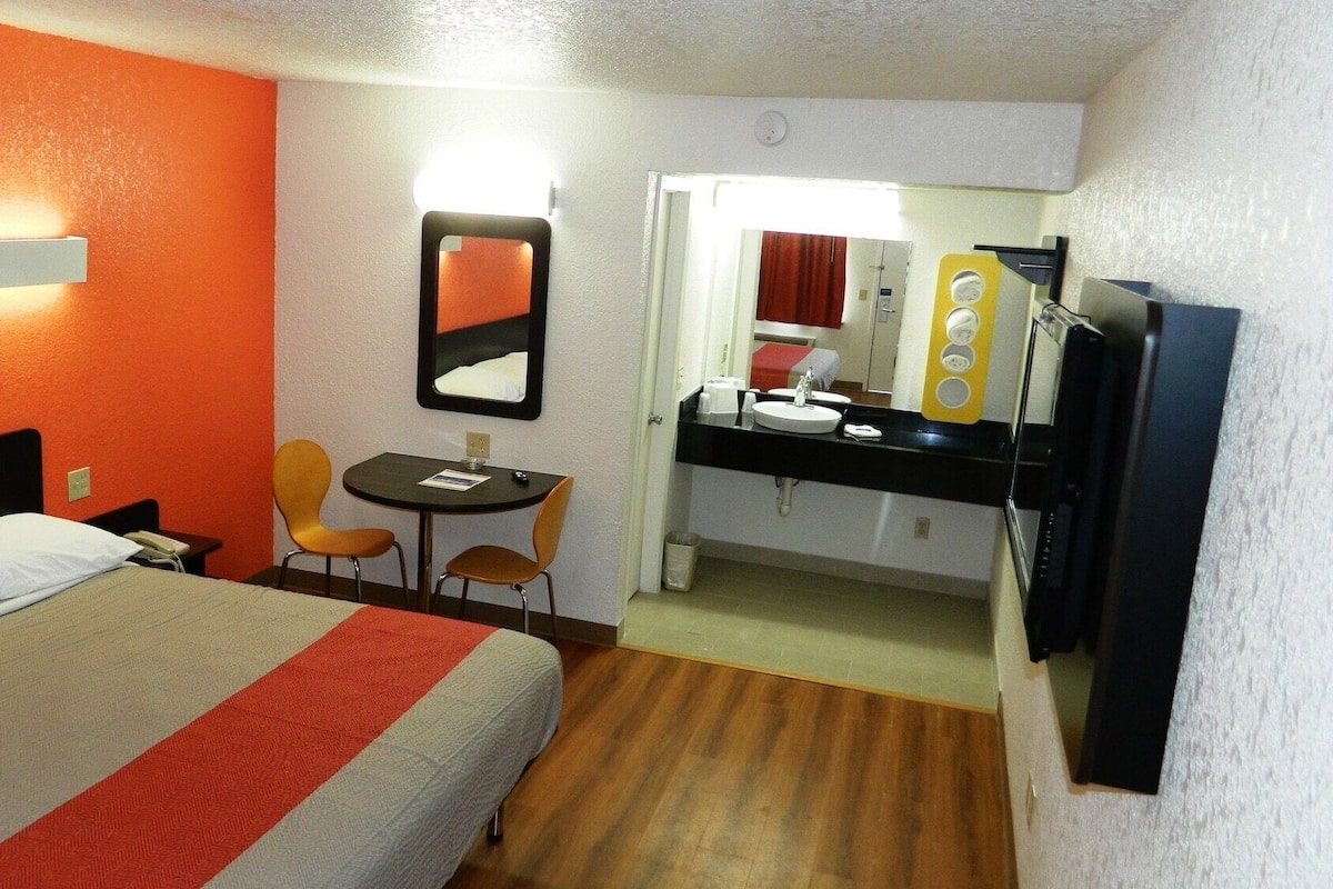Habitación de hotel con cama, escritorio, dos sillas, una mesita, espejo, lavamanos y televisor.