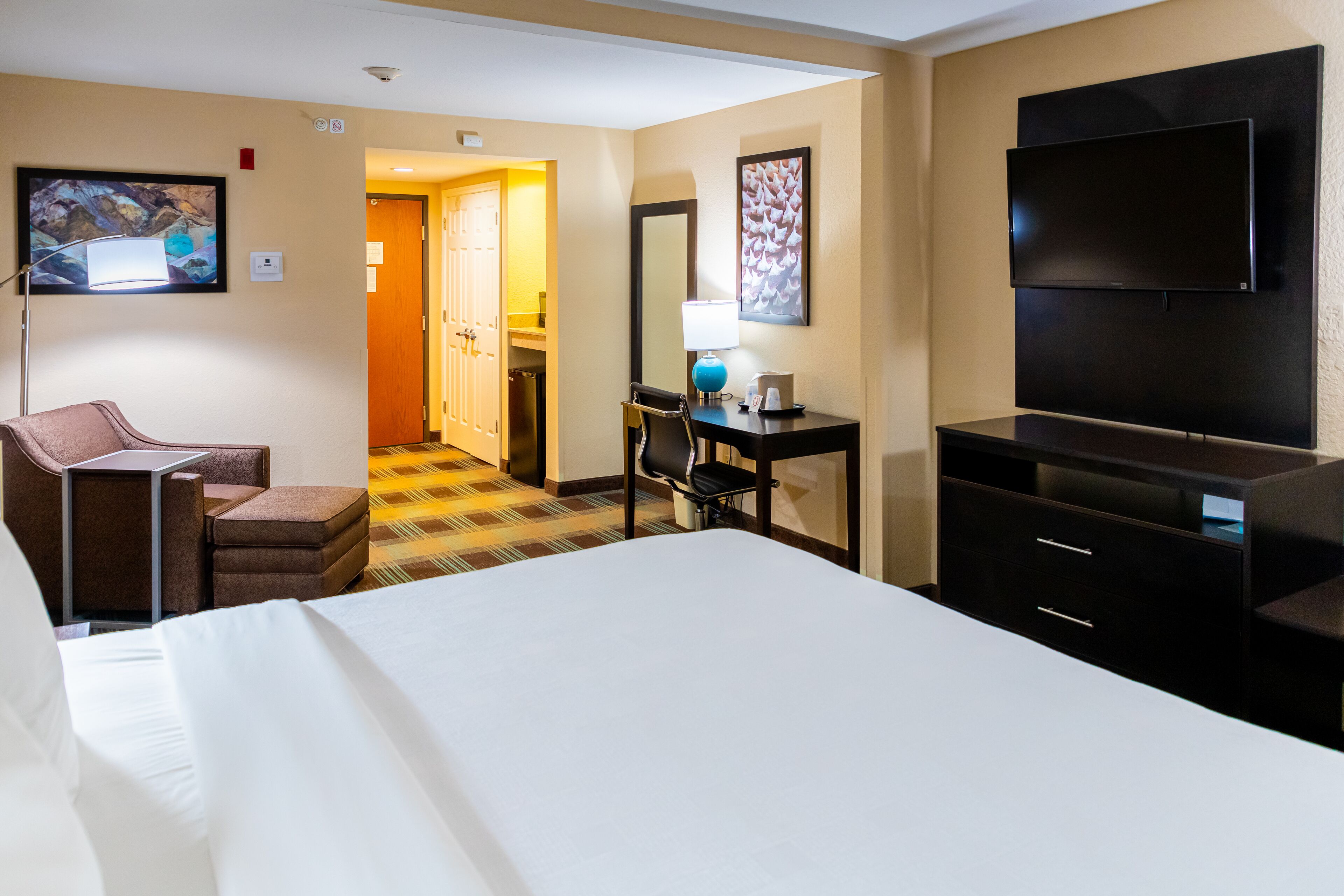 A hotel room with a bed, a desk with a chair, a television, and a door leading to another room.