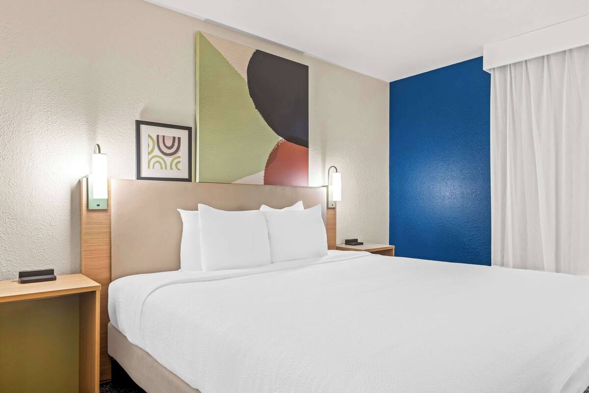 A hotel room with a large bed, two bedside tables, a nightstand, a wall-mounted lamp, and a framed picture.