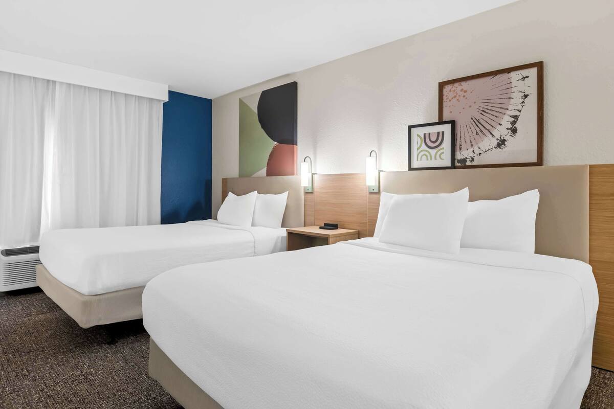 A hotel room with two beds, a large window with white curtains, a wall with abstract art, and a framed picture.