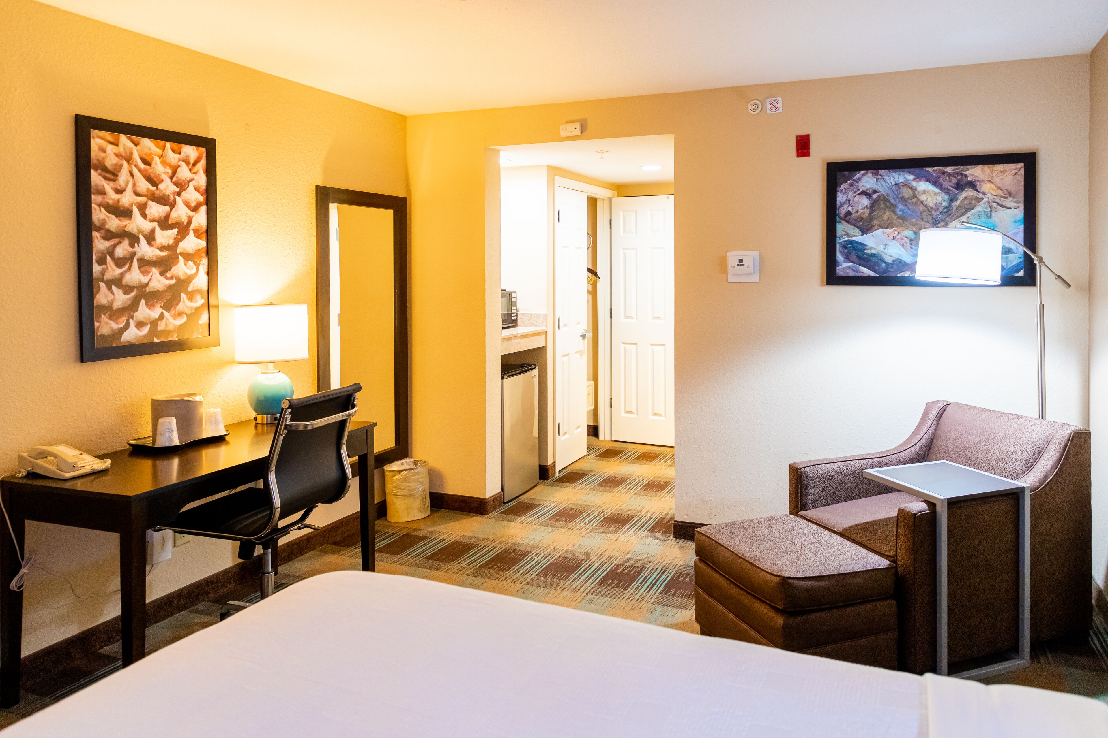 A hotel room with a bed, a desk with a telephone, a chair, a small table, a lamp, a trash bin, and a painting on the wall.