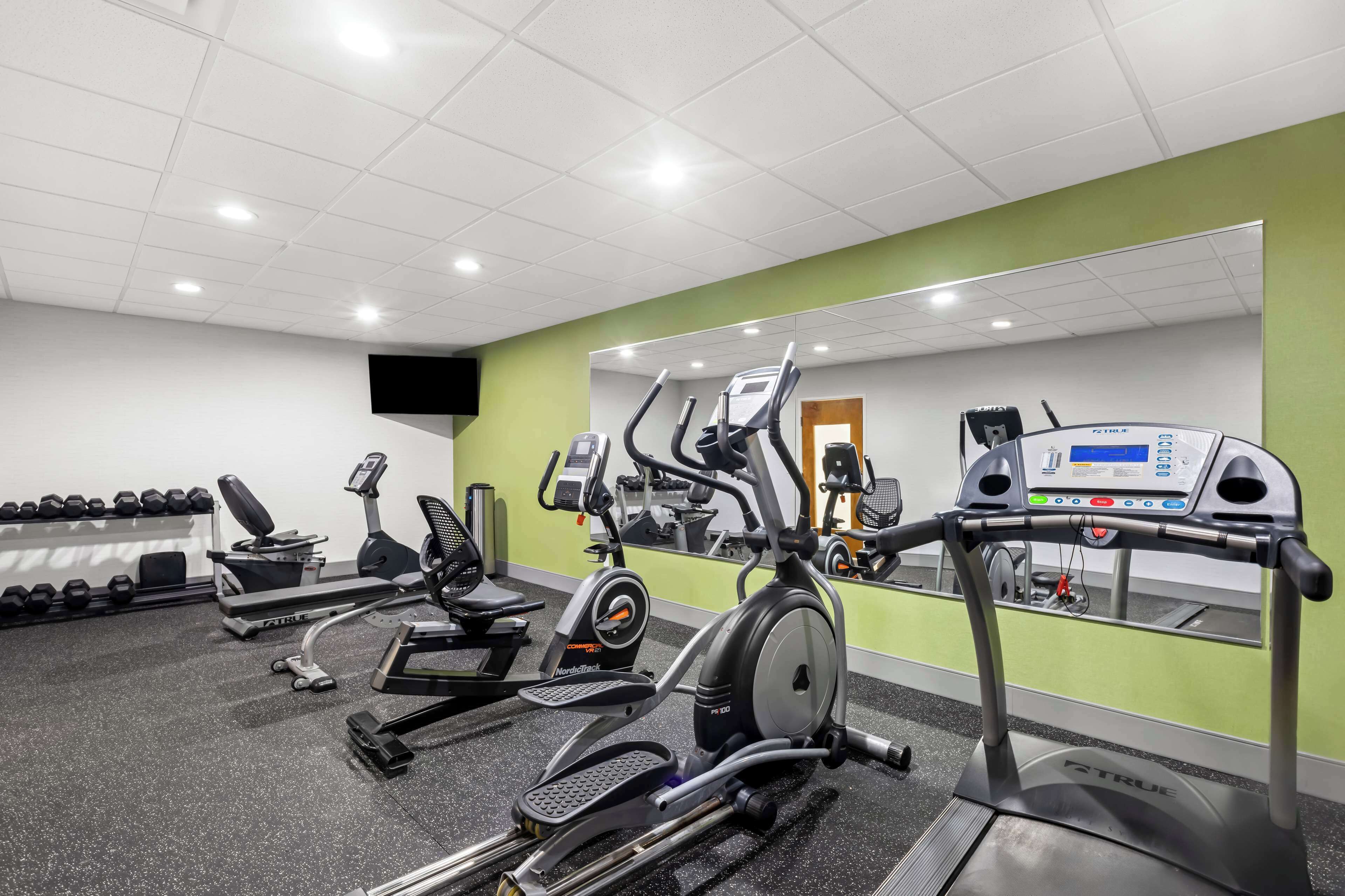 A gym with cardio machines, dumbbells, and a mirrored wall.