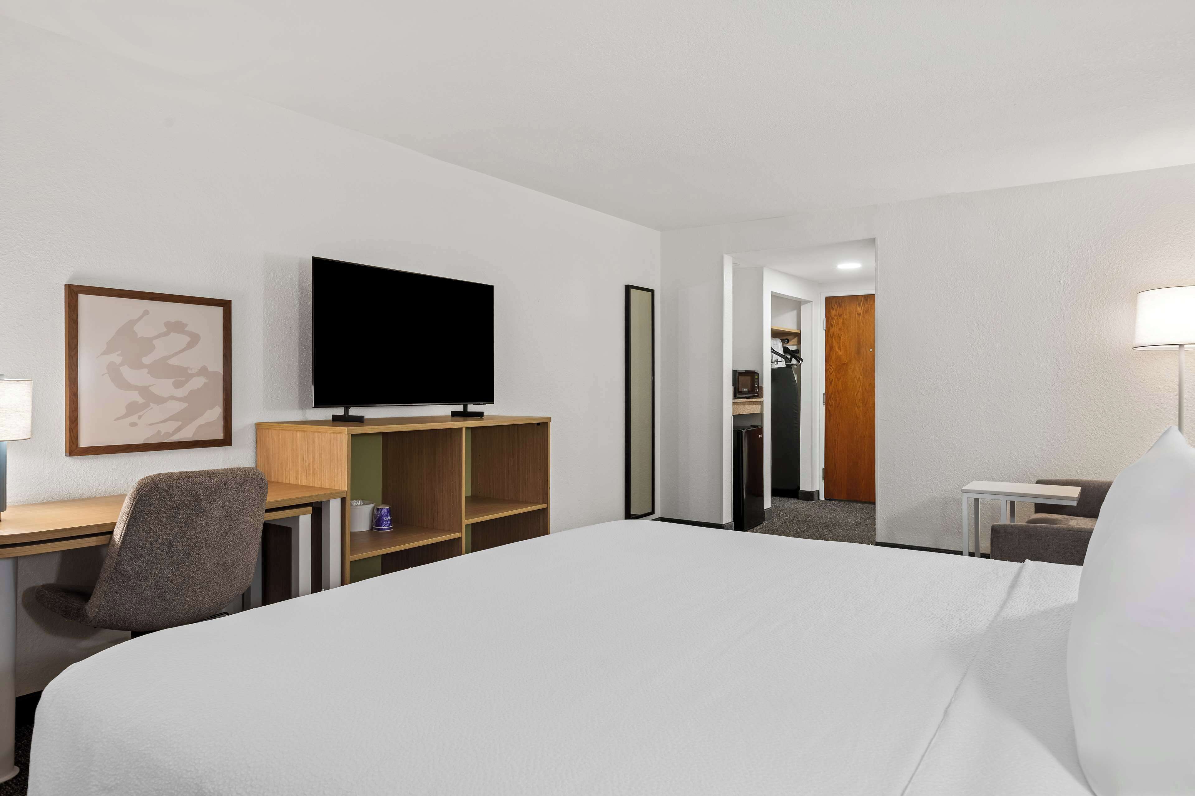 A hotel room with a large bed, a desk with a chair, a television, a small table, and a lamp.