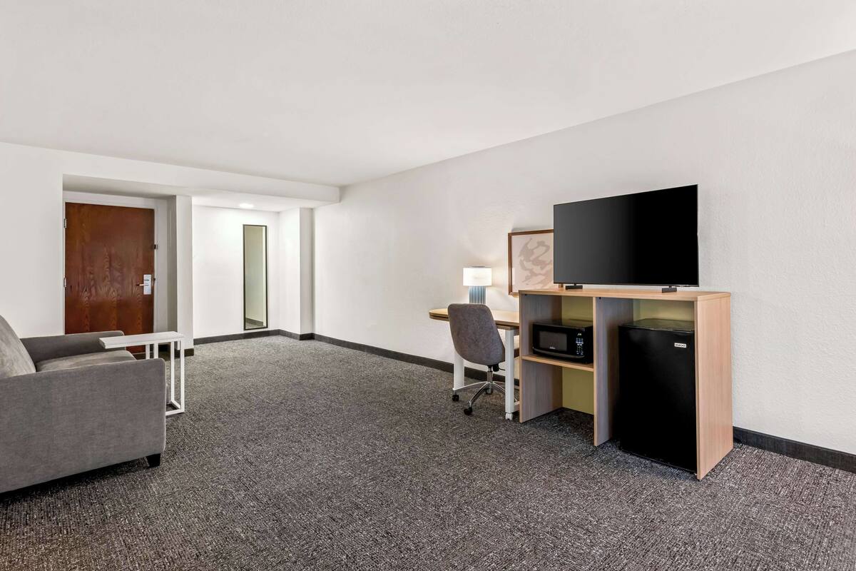 A hotel room with a grey sofa, a wooden desk with a TV and a small fridge, and a door leading to another room.