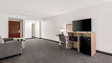 A hotel room with a grey sofa, a wooden desk with a TV and a small fridge, and a door leading to another room.