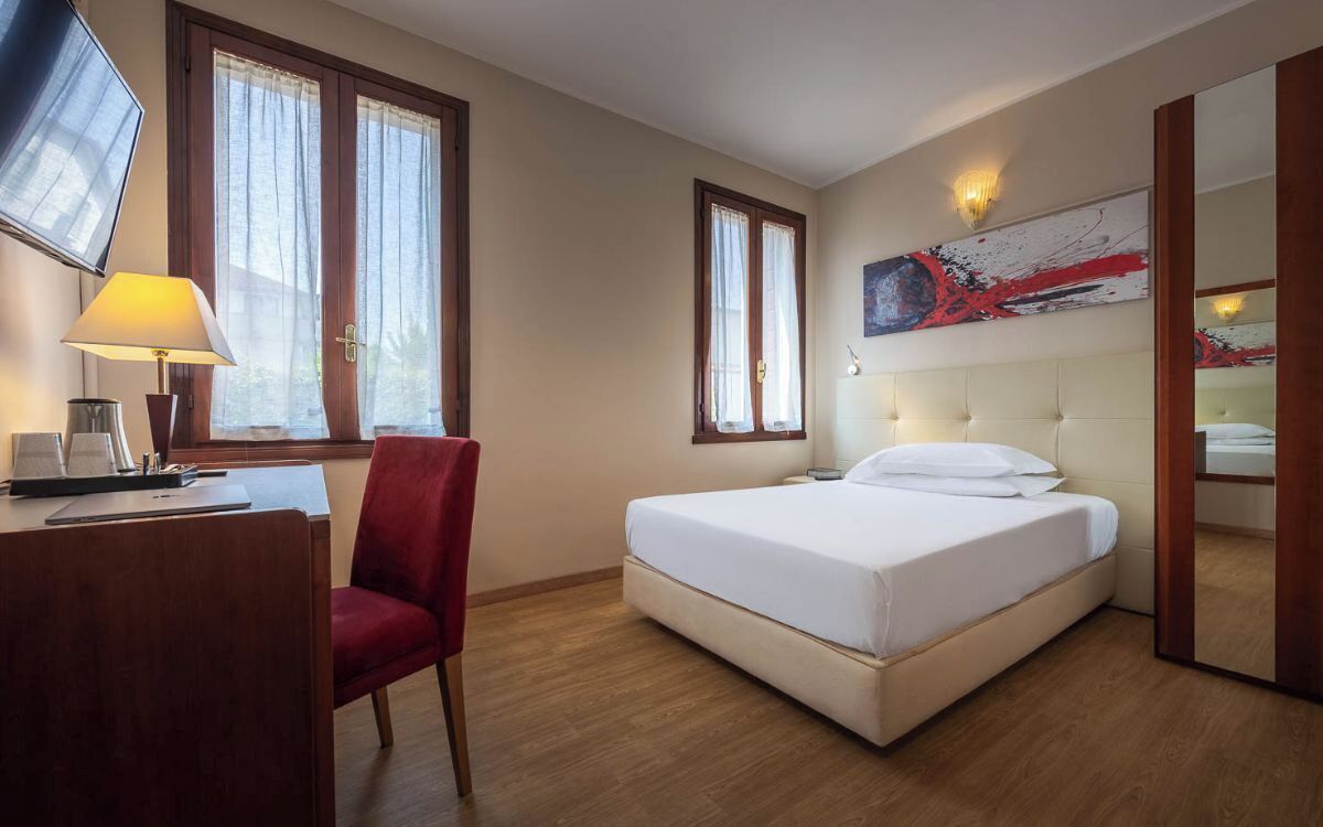 A hotel room with a bed, a desk with a lamp, a chair, and two windows.