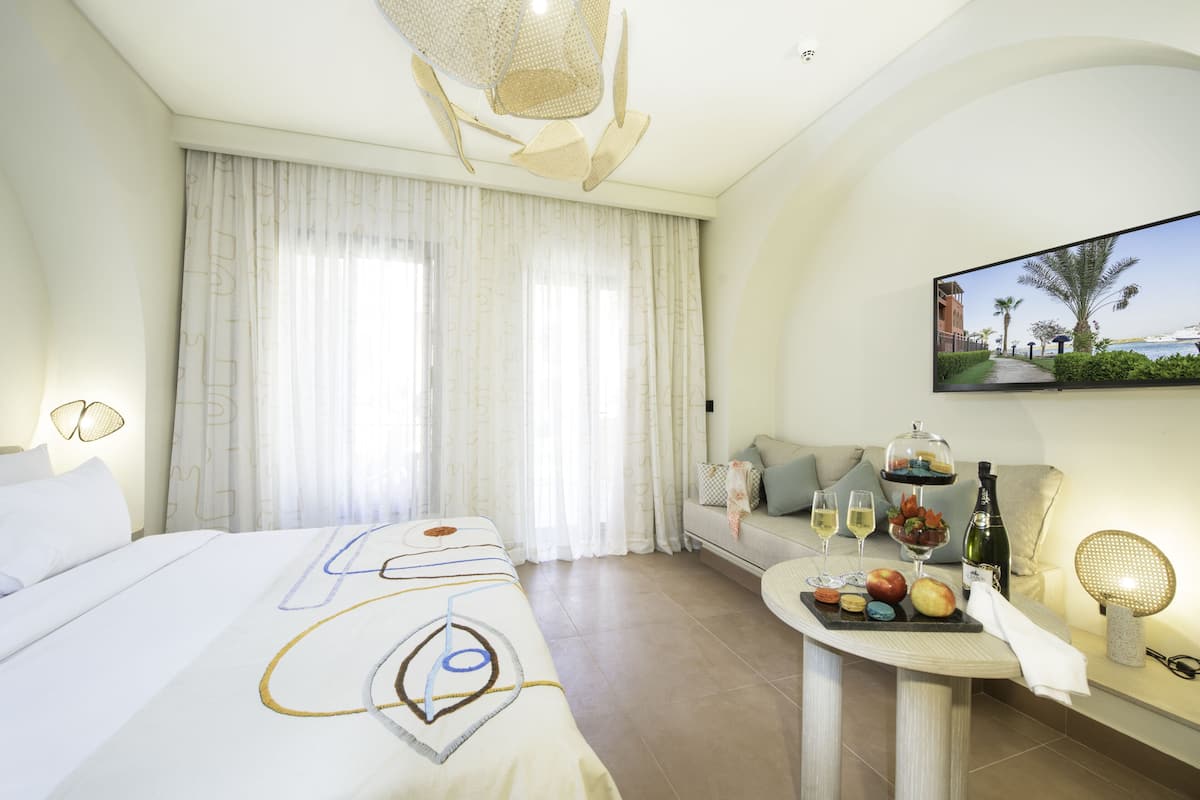 Hotelkamer met een bed, een zithoek, een tafel met eten en drinken, en een televisie waarop een strandscène te zien is.