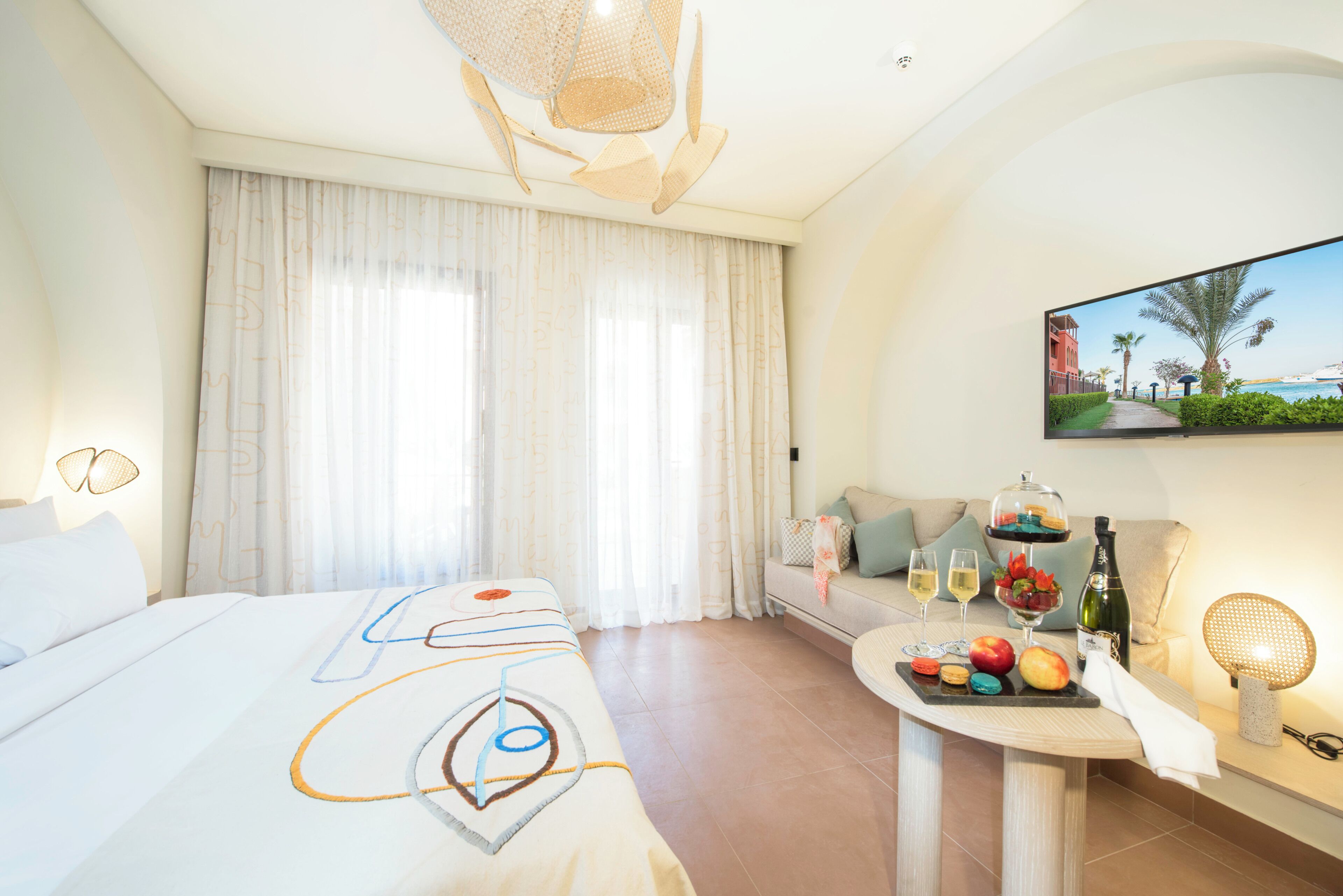 A hotel room with a bed, a seating area, a table with food and drinks, and a TV displaying a beach scene.