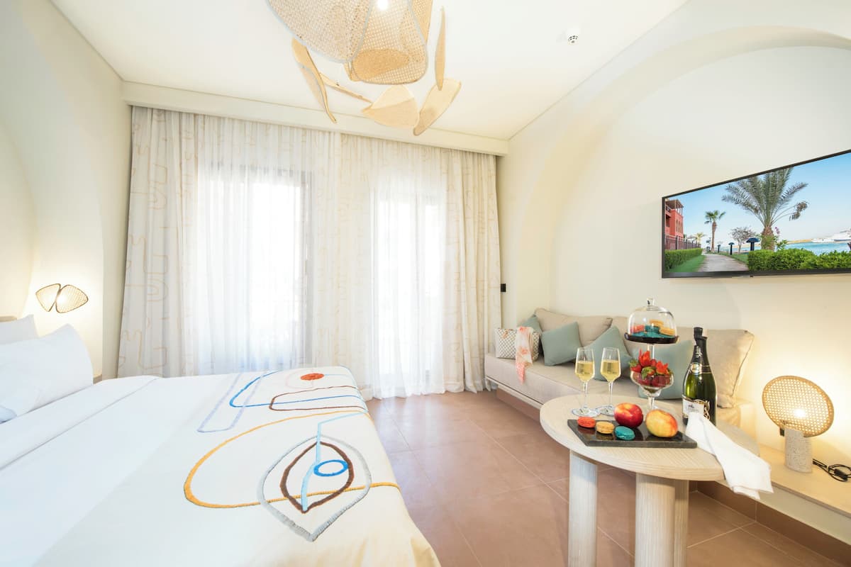 A hotel room with a bed, a seating area, a table with food and drinks, and a TV displaying a beach scene.