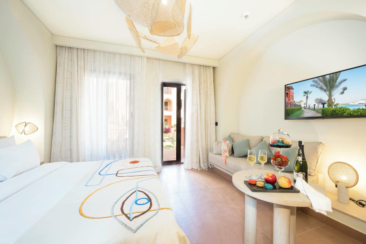 Hotelkamer met een bed, een bank, een eettafel met fruit en drankjes, en een televisie waarop een strand te zien is.