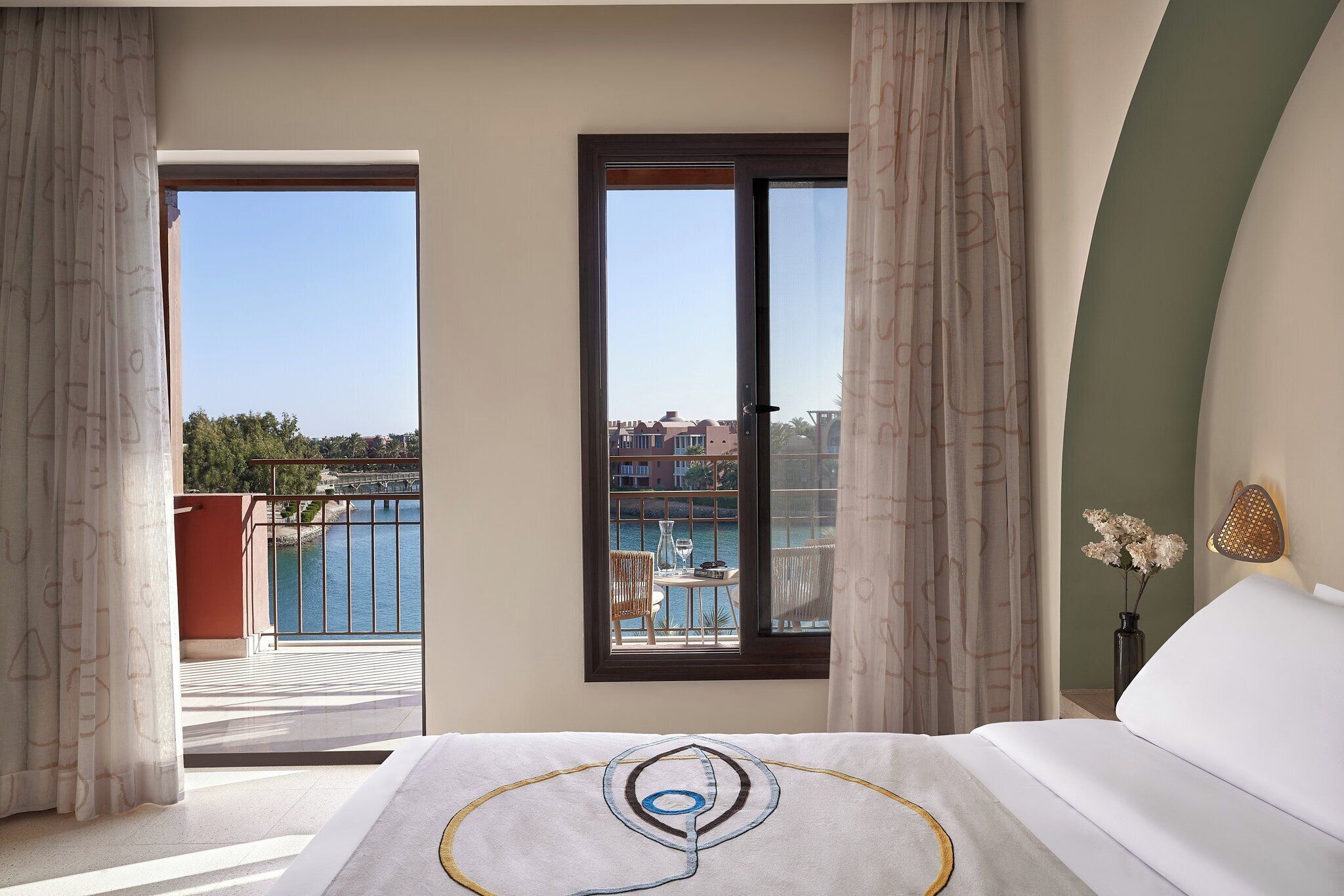 A hotel room with a bed, a view of a balcony with outdoor furniture, and a vase with flowers.