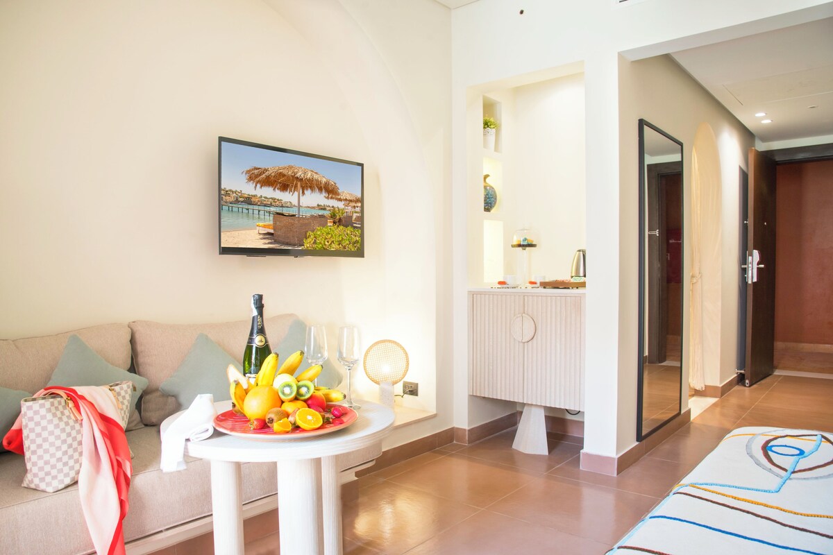 Une chambre d’hôtel moderne avec un canapé, une petite table sur laquelle se trouvent des fruits et une bouteille, un tableau représentant une scène de plage, et une salle de bain avec un miroir et un porte-serviettes.