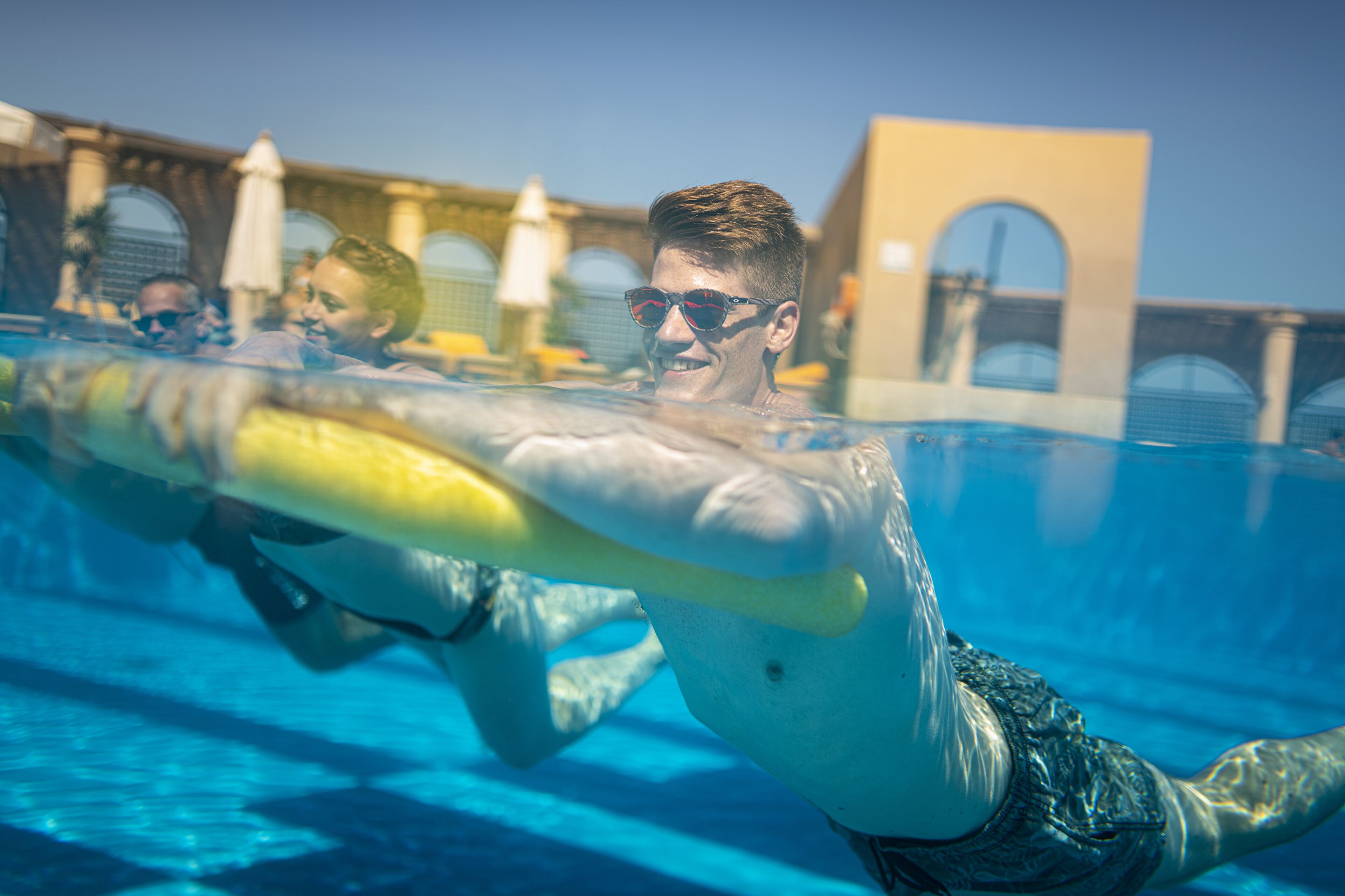 A swimming pool with clear blue water, a person swimming, and a building with arched windows in the background.
