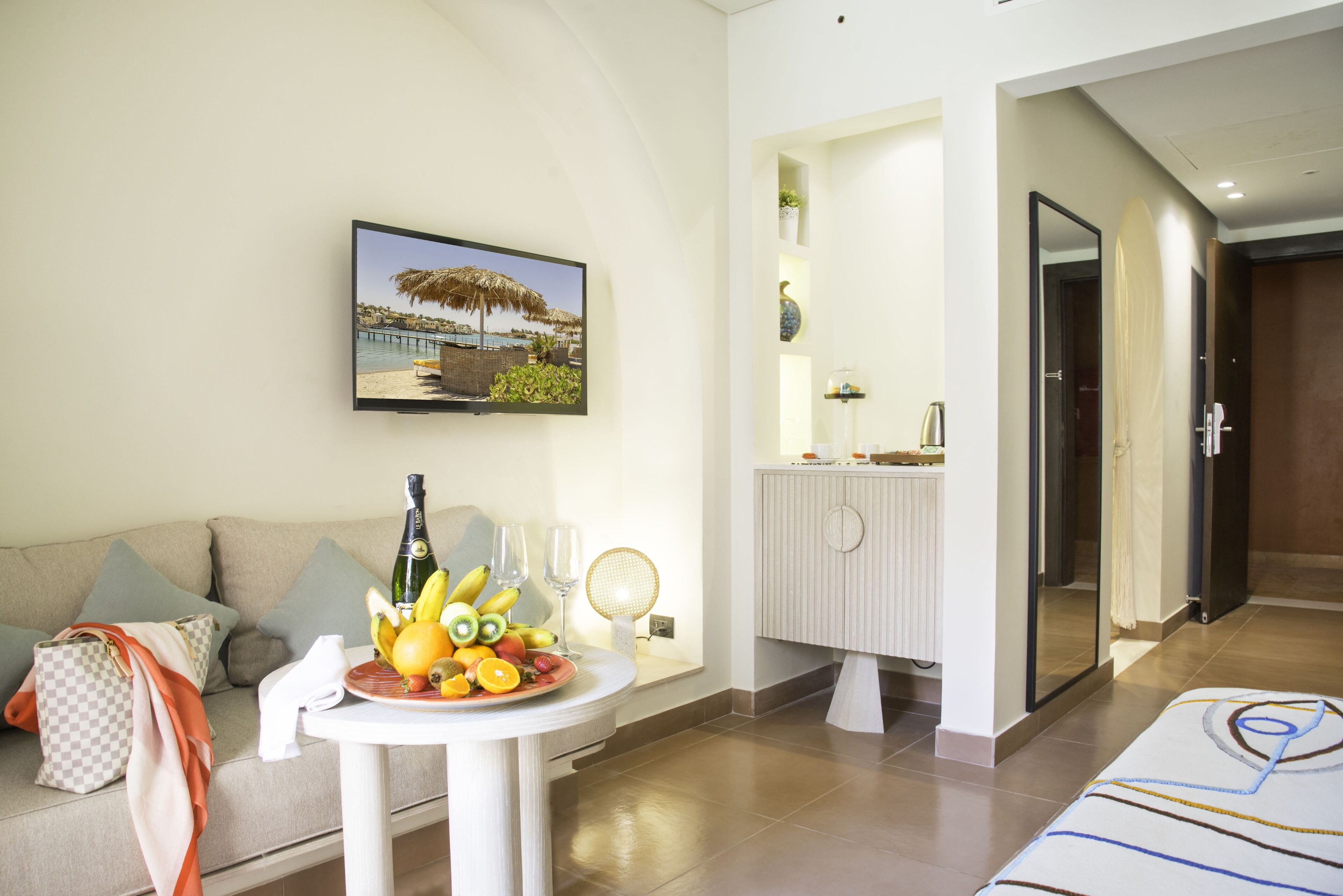 A modern hotel room with a sofa, a small table with fruit and a bottle, a painting of a beach scene, and a bathroom with a mirror and towel rack.