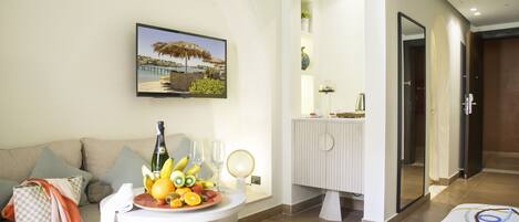 A modern hotel room with a sofa, a small table with fruit and a bottle, a painting of a beach scene, and a bathroom with a mirror and towel rack.