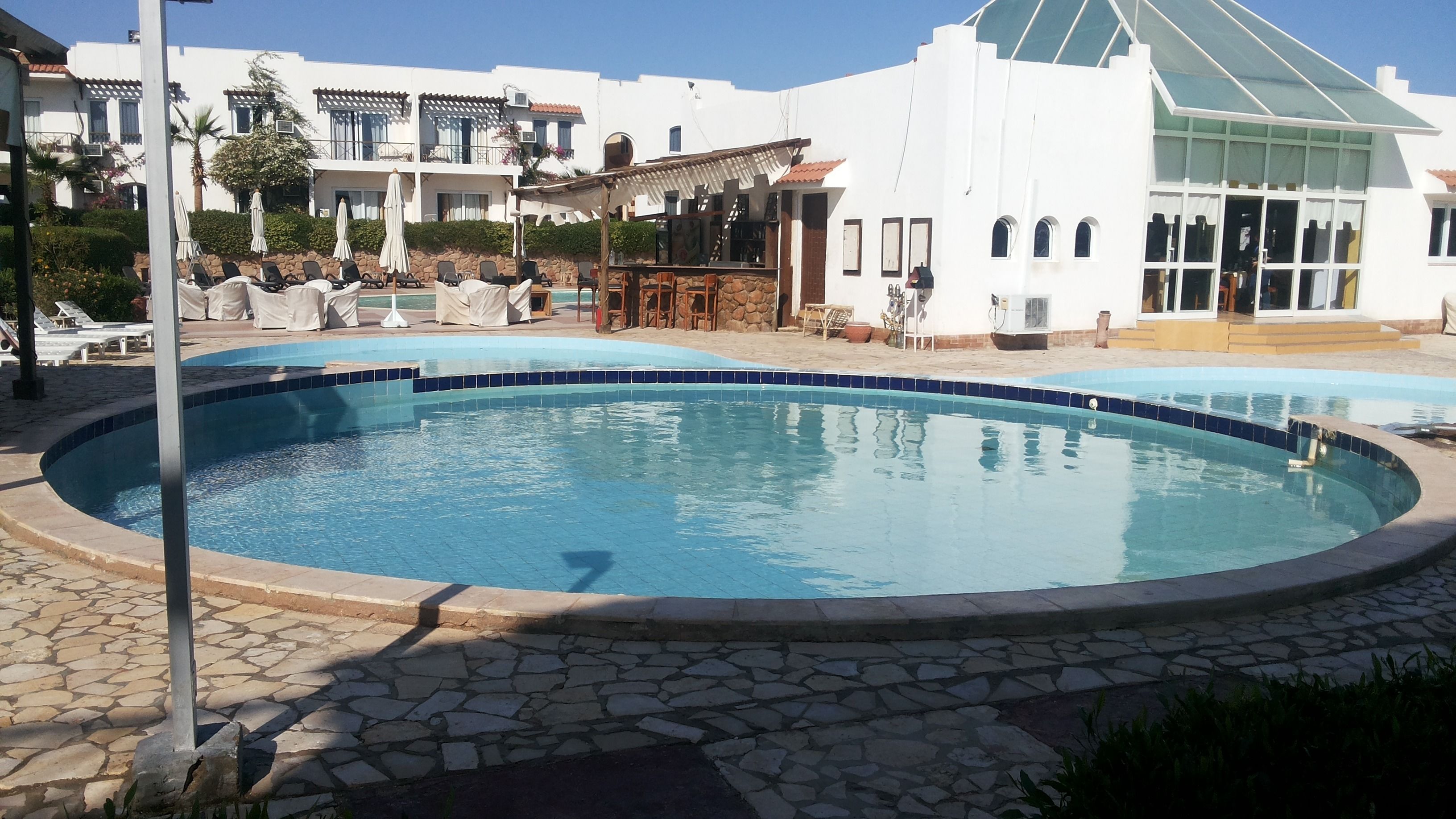 A swimming pool with lounge chairs and umbrellas, surrounded by stone tiles and a building with a white facade and arched windows.
