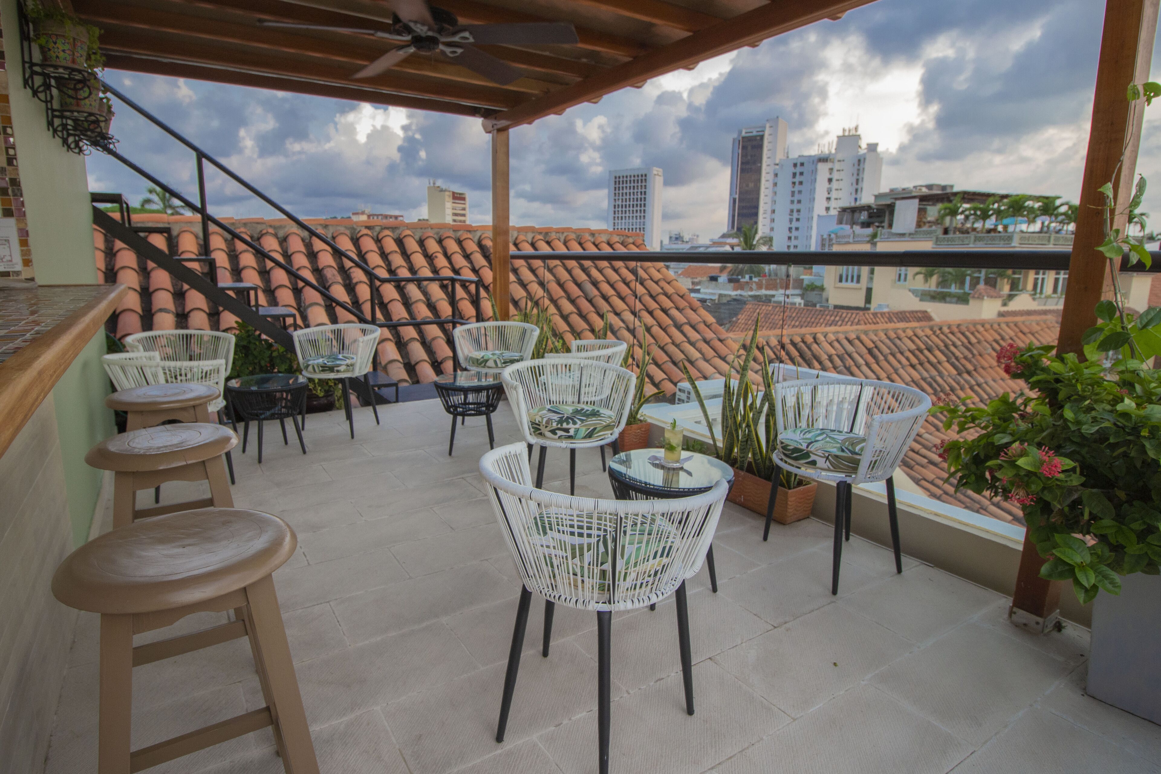 A rooftop terrace with wicker chairs and stools, a view of city buildings, and a tiled roof.
