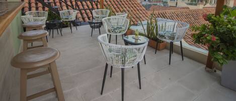 A rooftop terrace with wicker chairs and stools, a view of city buildings, and a tiled roof.