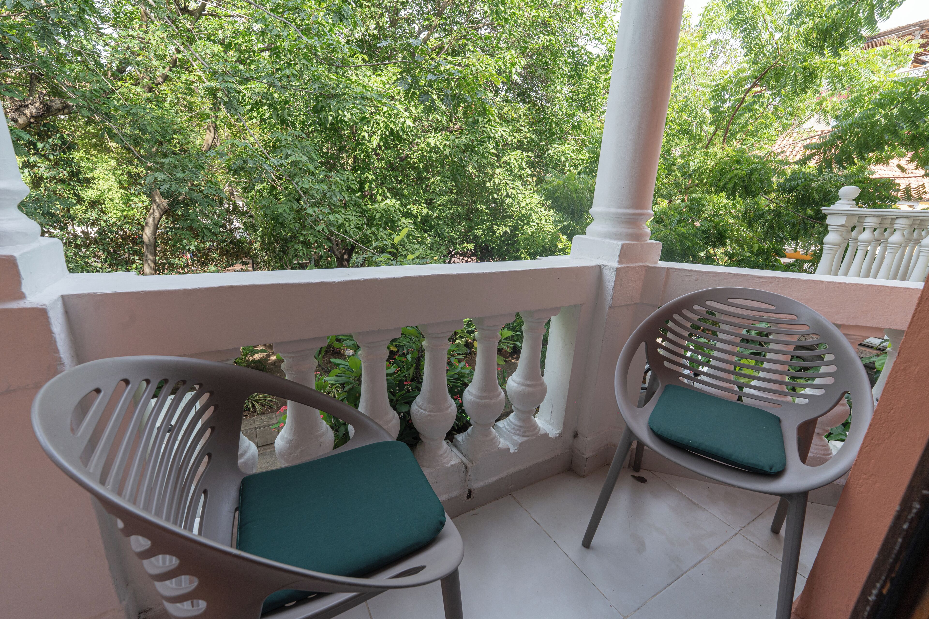 A balcony with two white plastic chairs with green cushions, a white balustrade, and lush green trees in the background.
