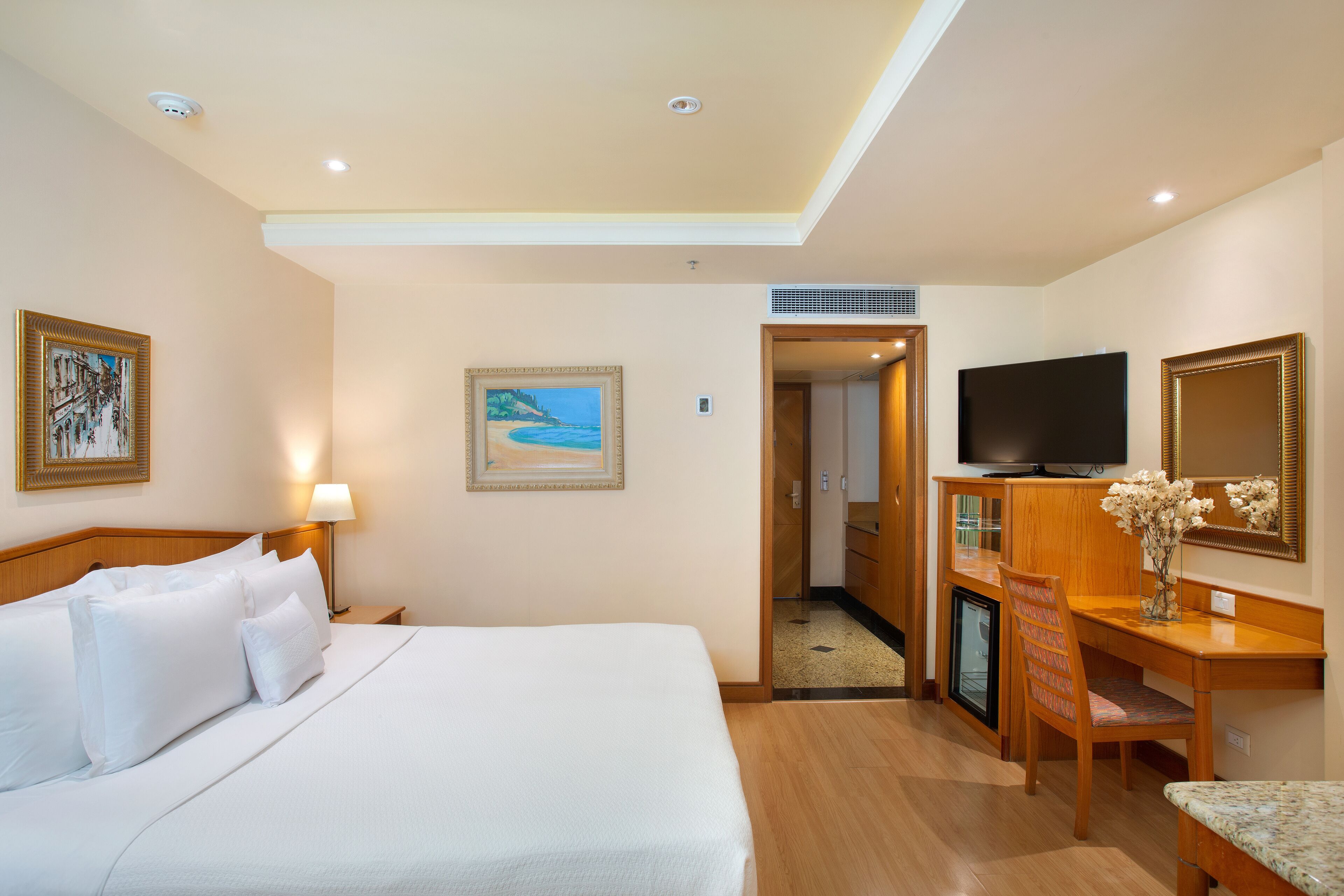 A hotel room with a bed, a desk, a television, and a painting on the wall.
