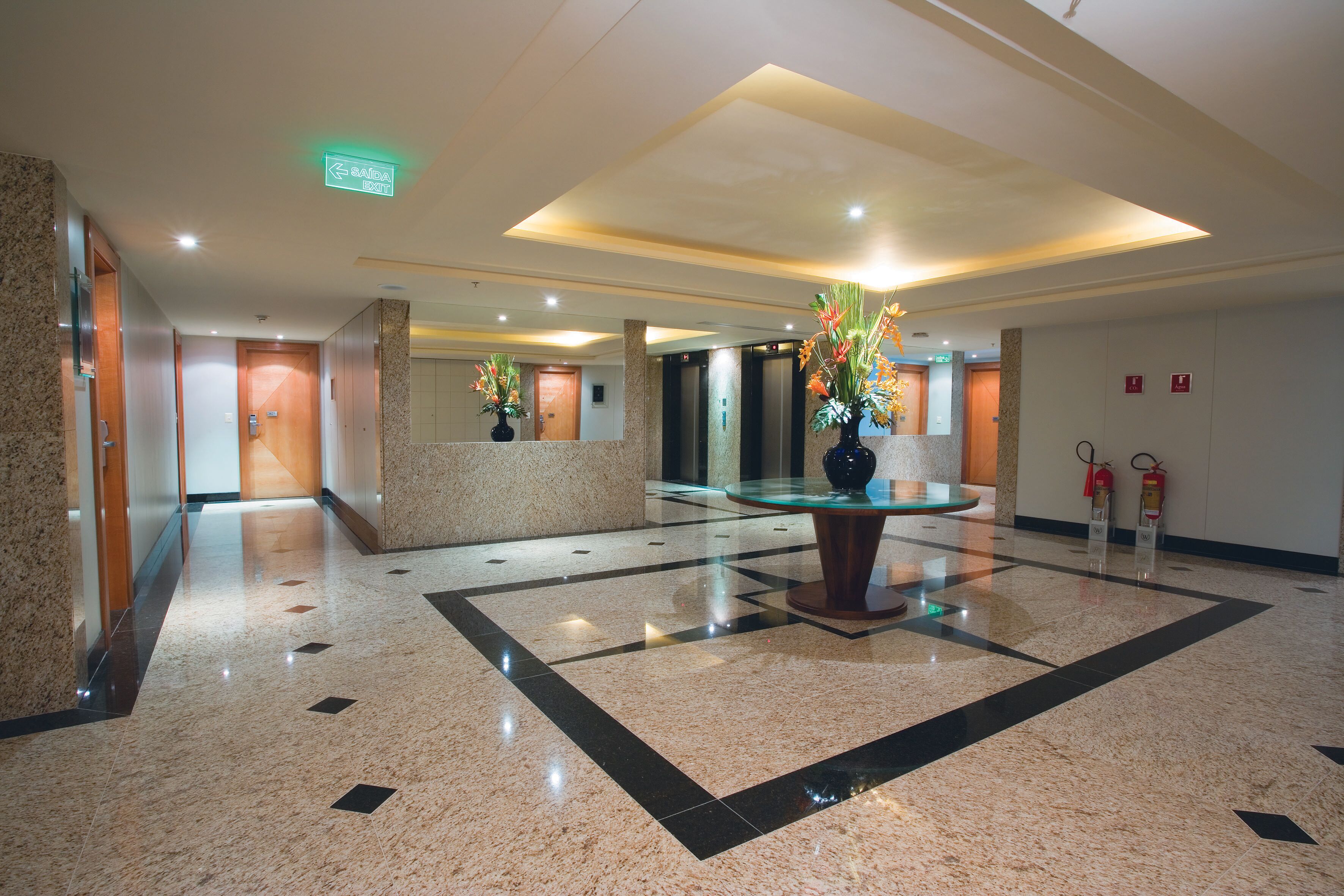 A lobby with a glass reception desk, a central decorative plant, and a fire extinguisher on the wall.