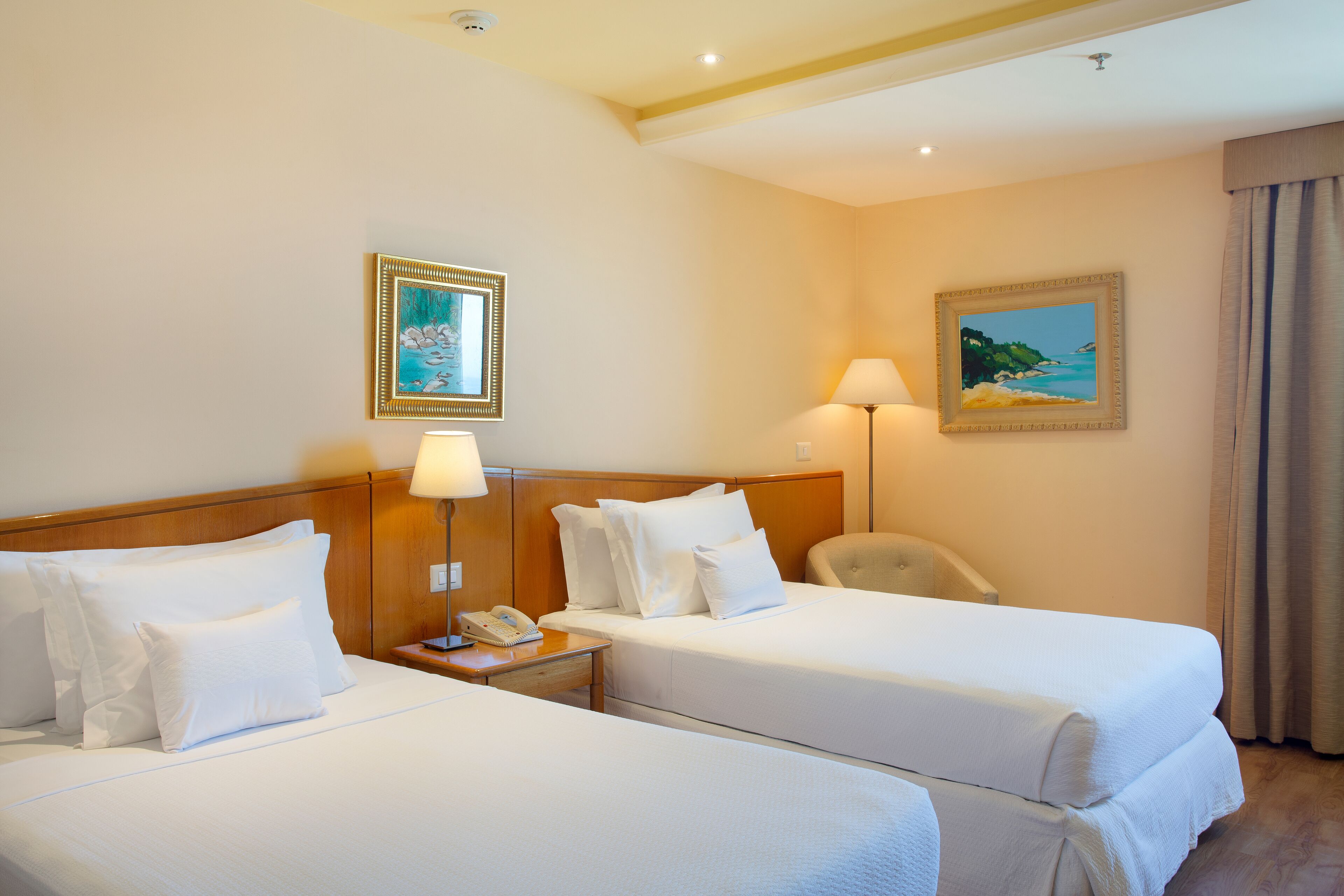 Habitación de hotel con dos camas, una mesita de noche con lámpara, un teléfono y dos cuadros en la pared.