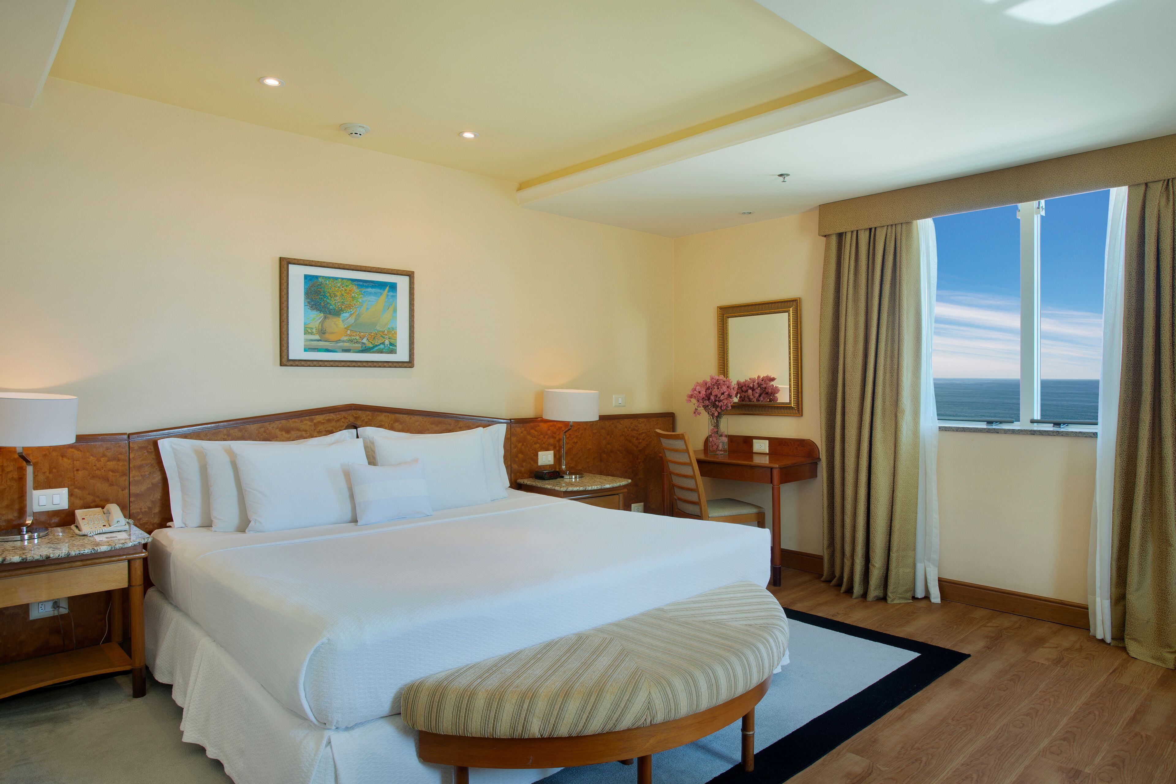 Una habitación de hotel con una cama grande, un escritorio, una silla, una mesita con un jarrón y una ventana con cortinas.