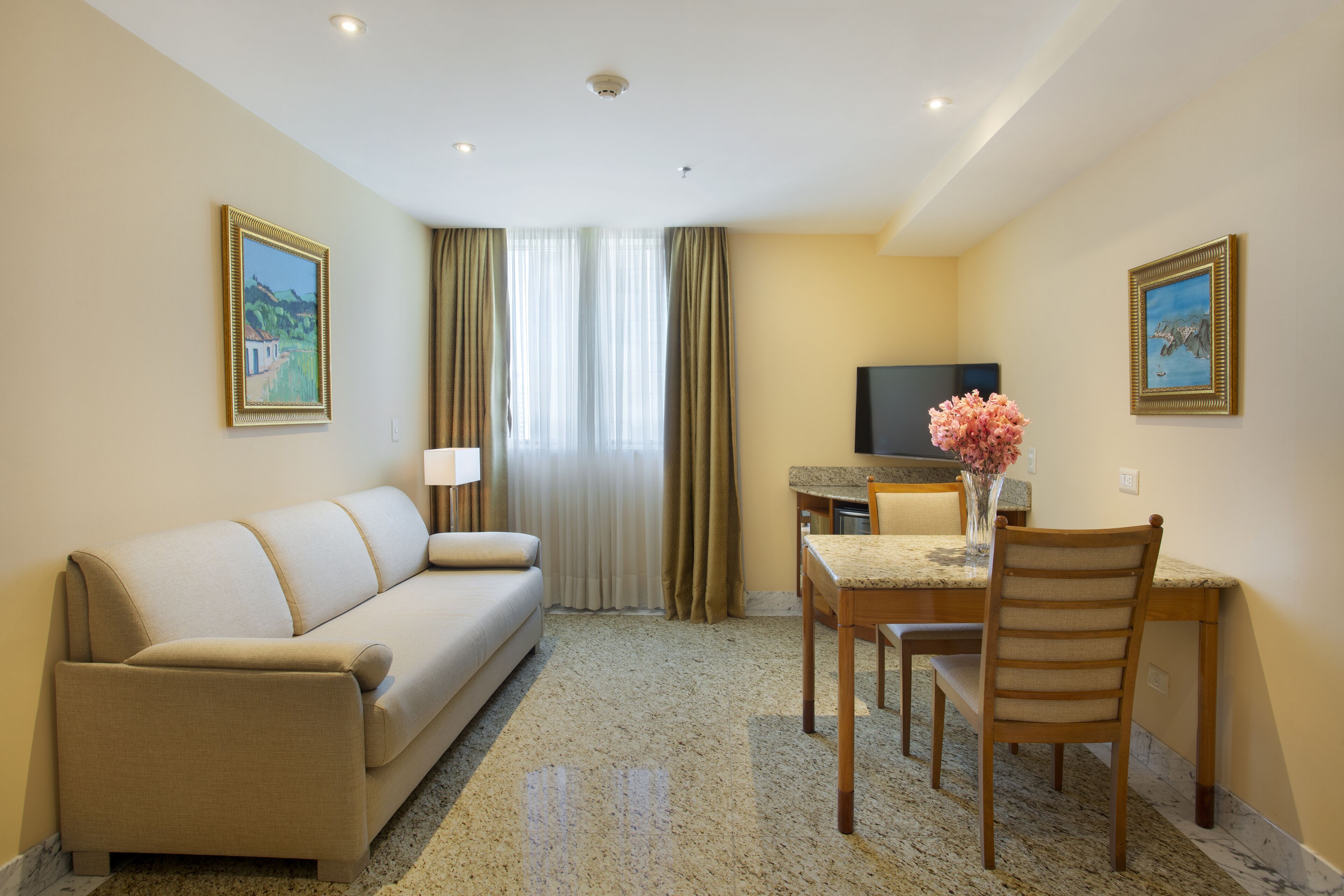 A hotel room with a sofa, a dining table with chairs, a TV, and a vase with flowers.