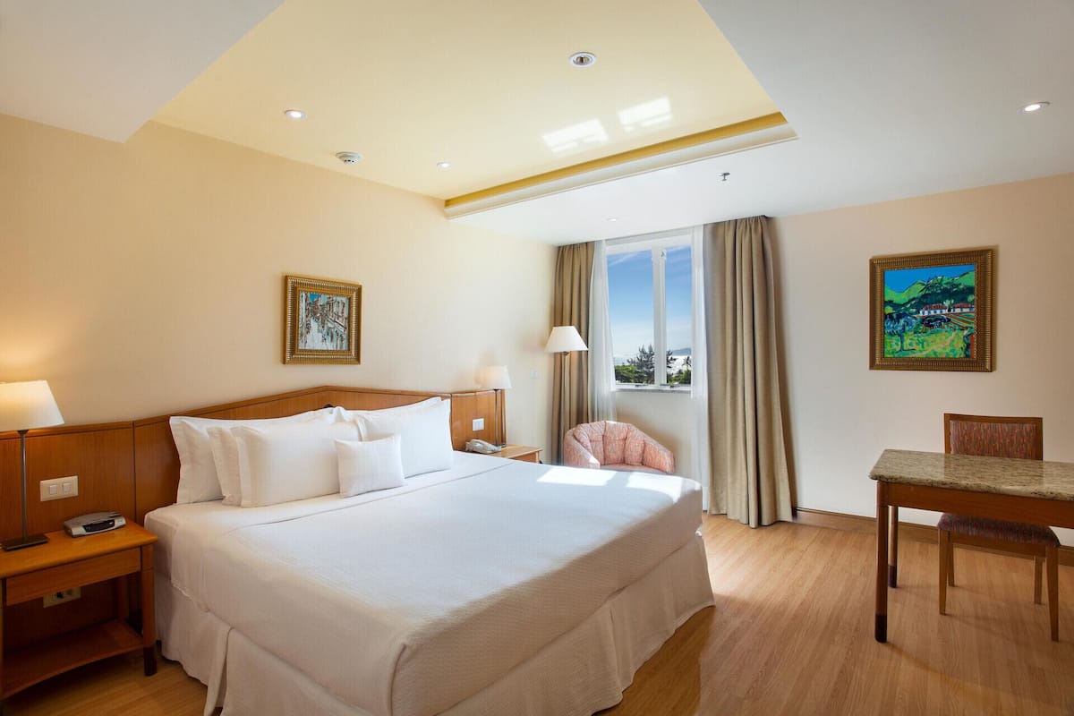 Habitación de hotel con una cama grande, mesitas de noche, un escritorio y una silla. Hay dos cuadros en las paredes, una ventana con cortinas y un techo con iluminación empotrada.