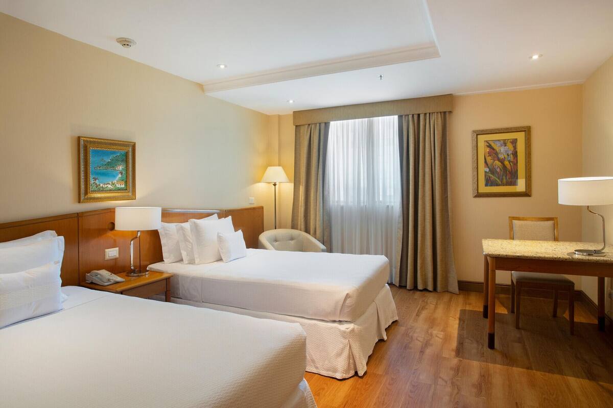 Habitación de hotel con dos camas, un escritorio, una silla, una lámpara, un cuadro y una ventana con cortinas.