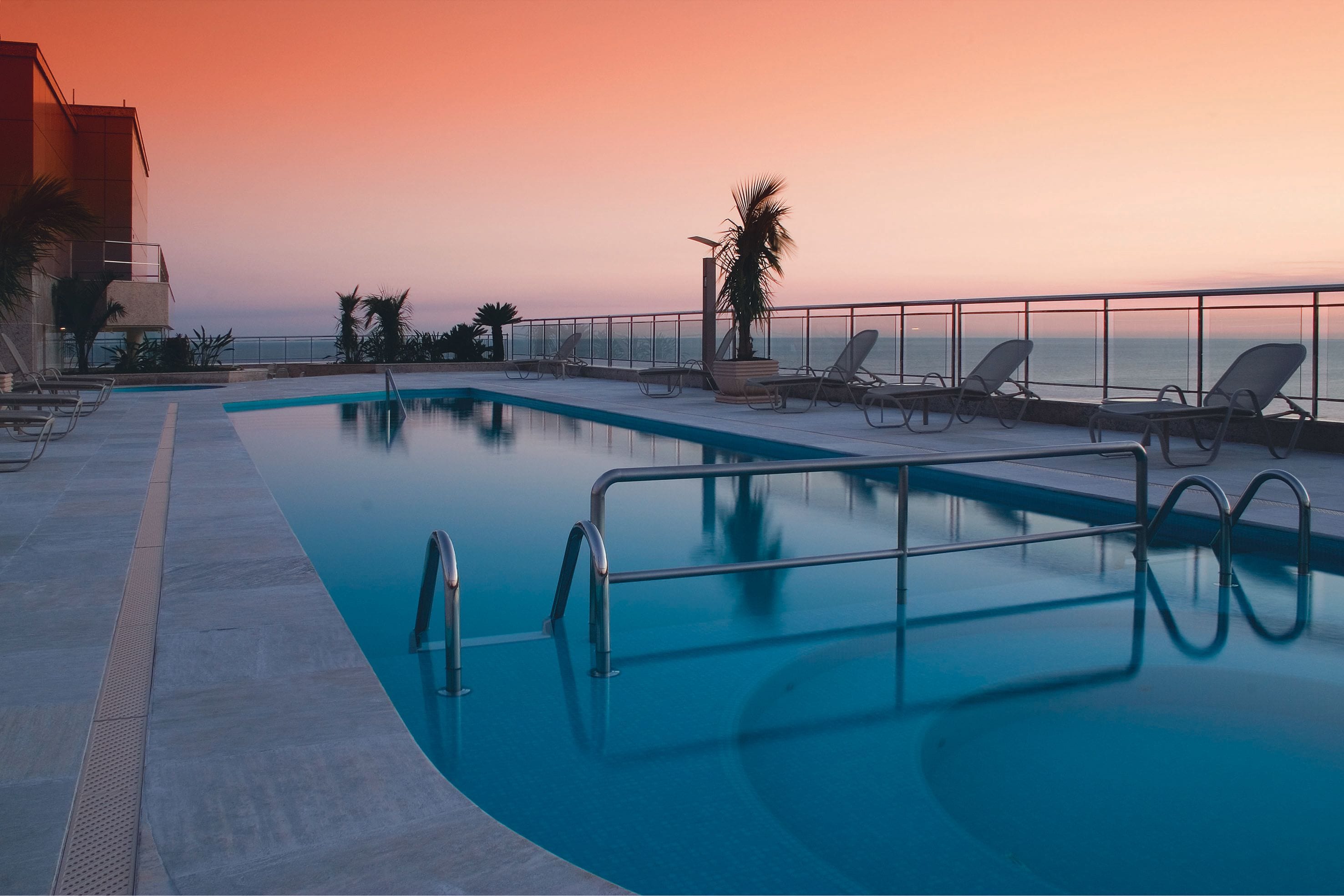 Une piscine avec des chaises longues et un palmier, donnant sur une étendue d’eau au coucher du soleil.