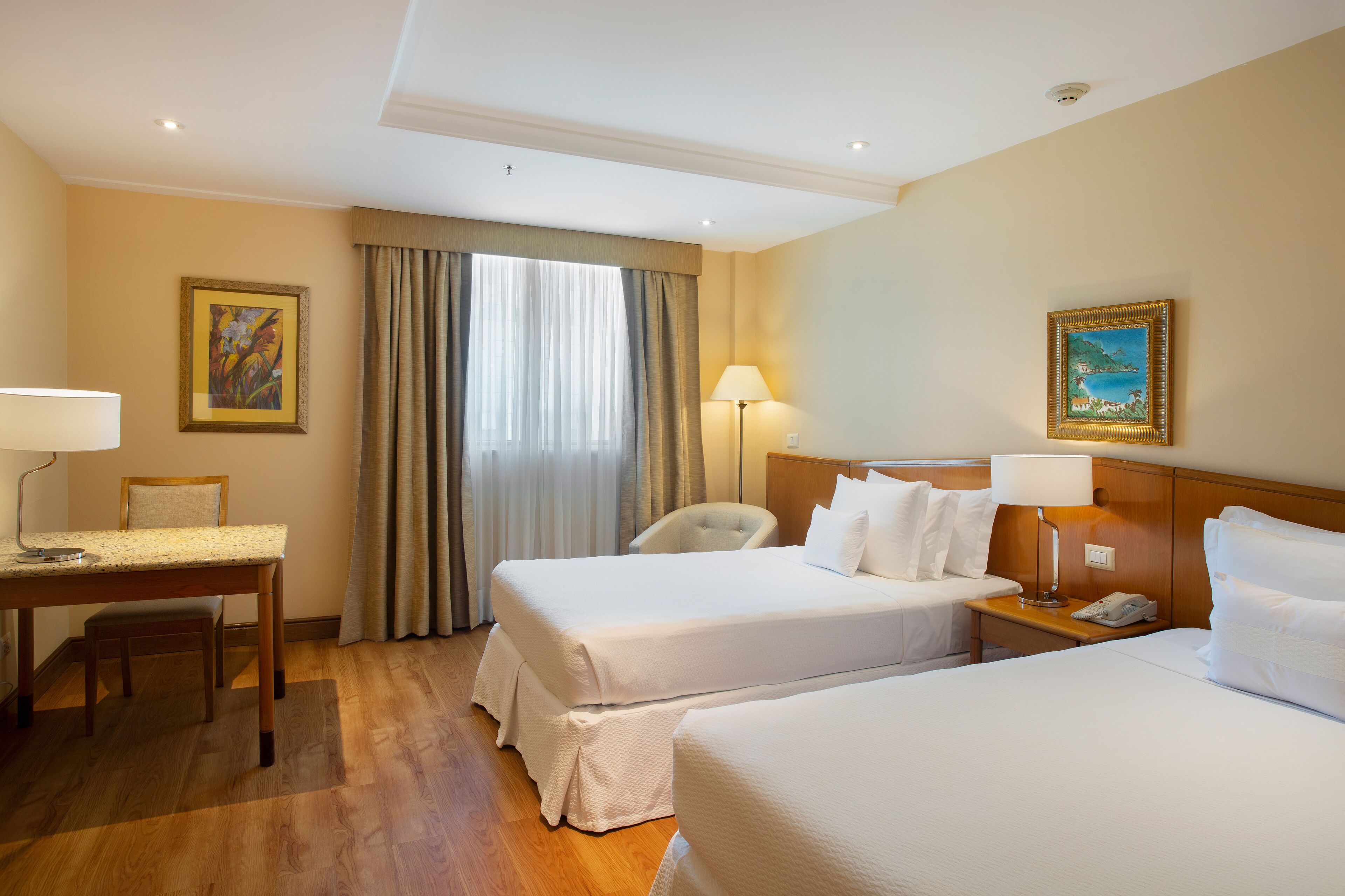 Quarto de hotel com duas camas, uma escrivaninha, uma cadeira, um abajur e um quadro na parede.